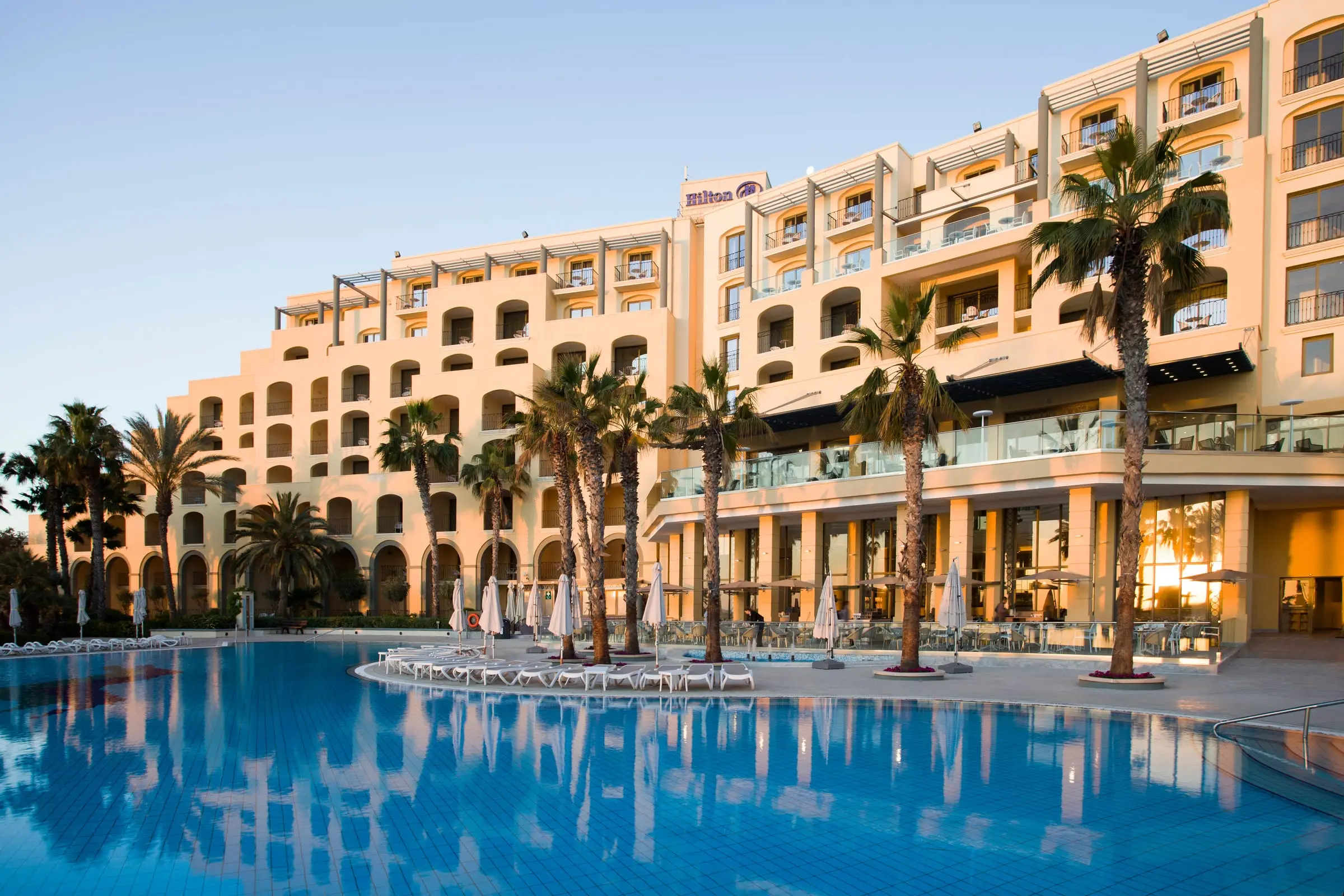Hilton Malta in evening sun, its terraces and palms reflected in the main pool