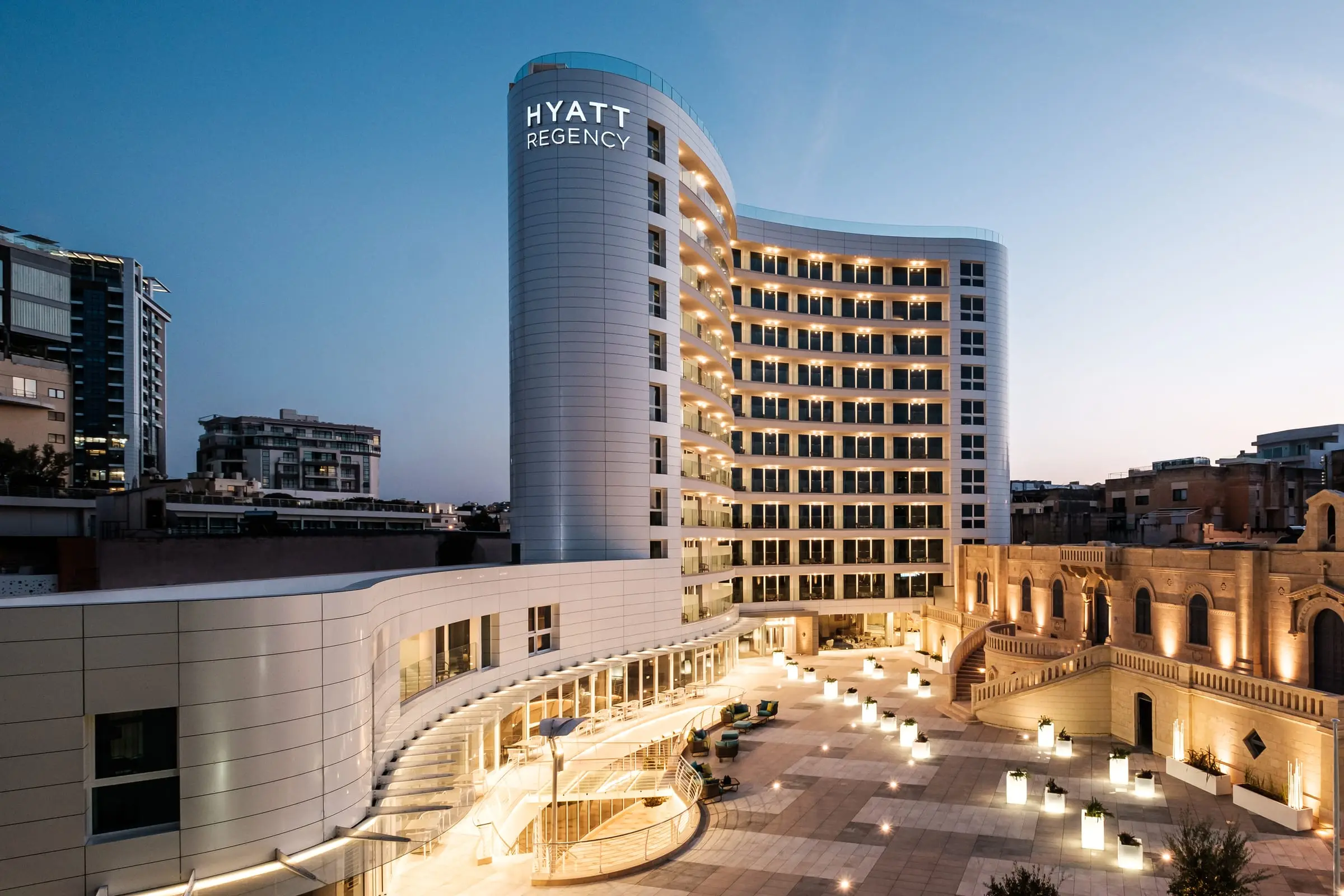 Hyatt Regency Malta at blue hour, its curved tower above the lit piazza and the Santa Rita priory