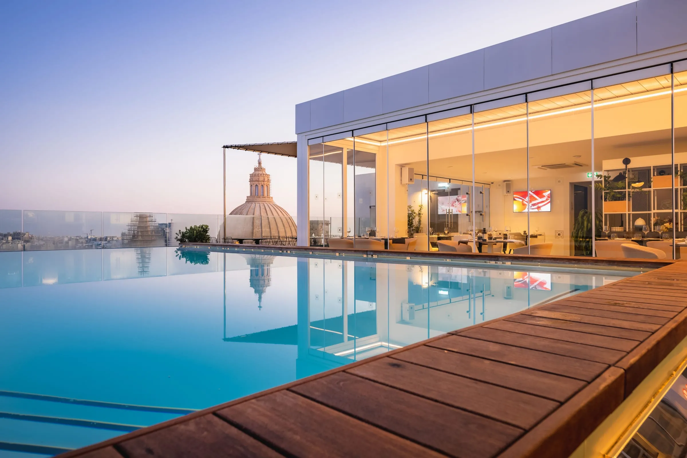 The rooftop pool at dusk beside the glass-walled restaurant, with a Valletta church dome beyond