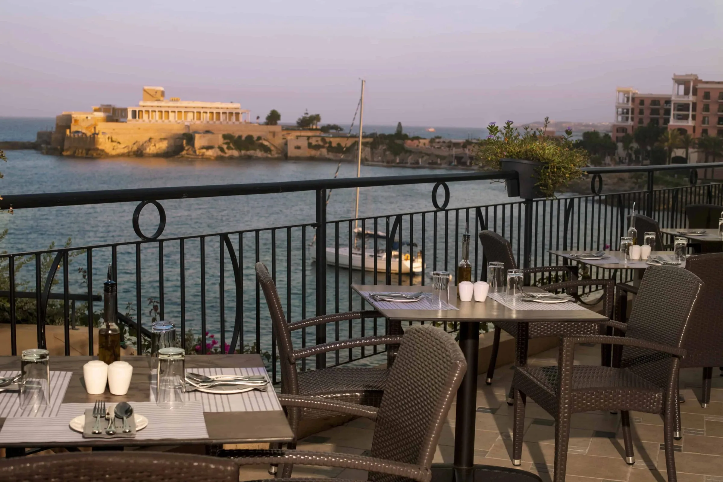 Restaurant tables on a terrace above St George's Bay in evening light, facing the Dragonara Palace