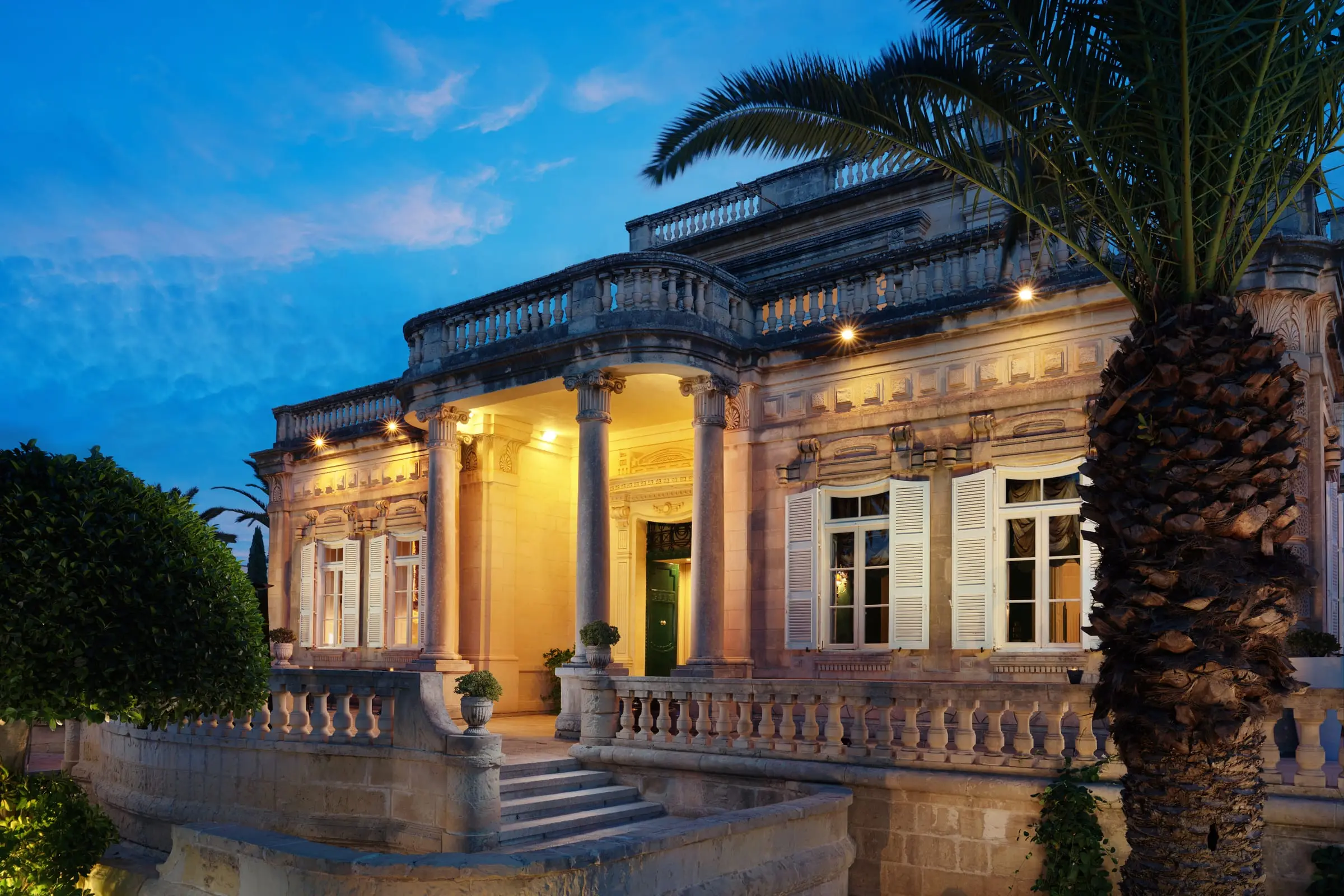 Villa Corinthia at dusk, its columned porch lit, with a palm in front
