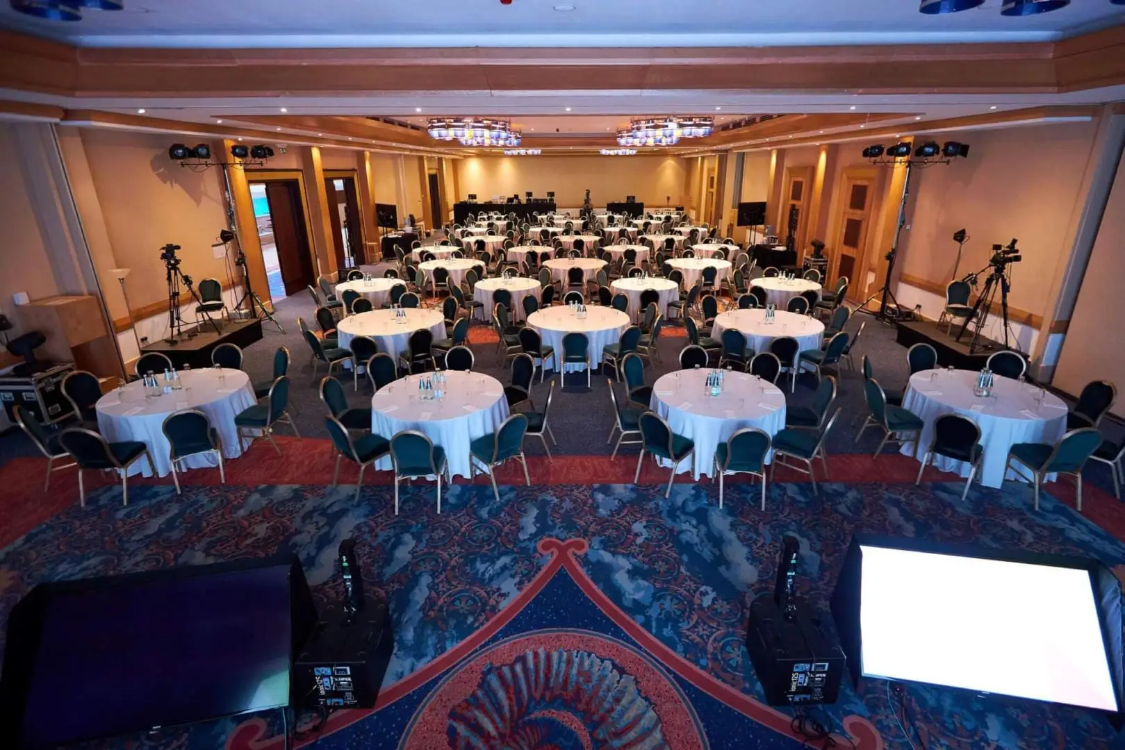 A ballroom set with round tables for a conference, with cameras and stage lighting in place