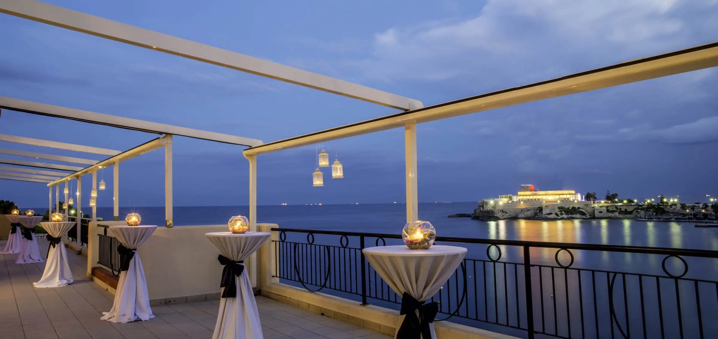 A seafront terrace set with cocktail tables at dusk, the lit Dragonara Palace across the bay