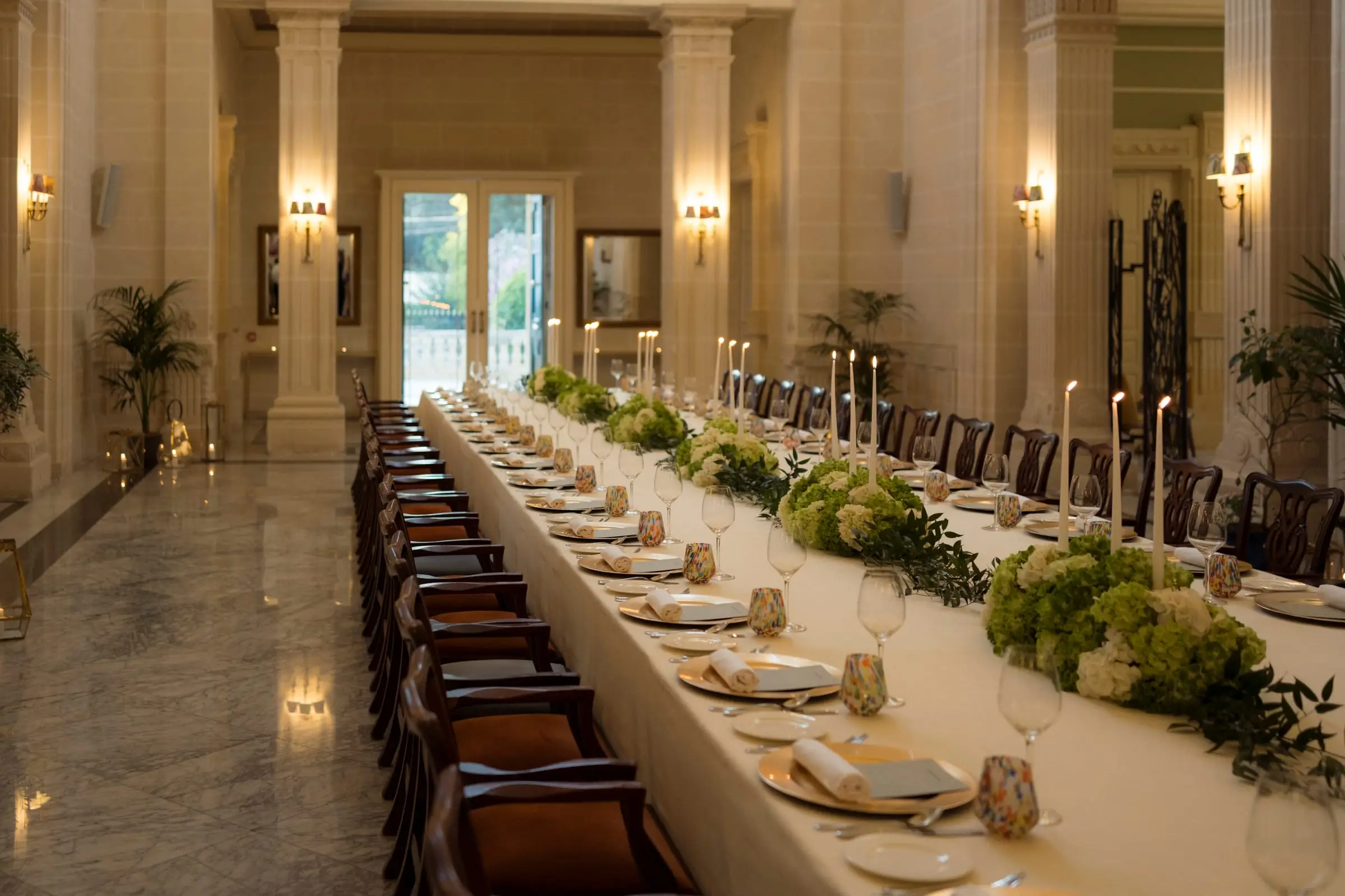 A long table laid for dinner in a columned hall, with candles and hydrangeas