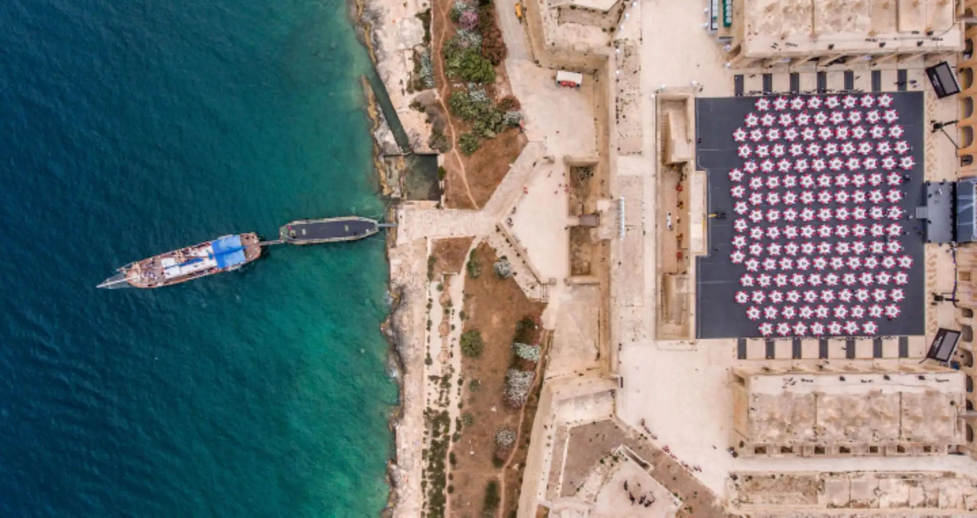A gala dinner laid out inside a fort on the water, seen from the air, a boat moored at the jetty