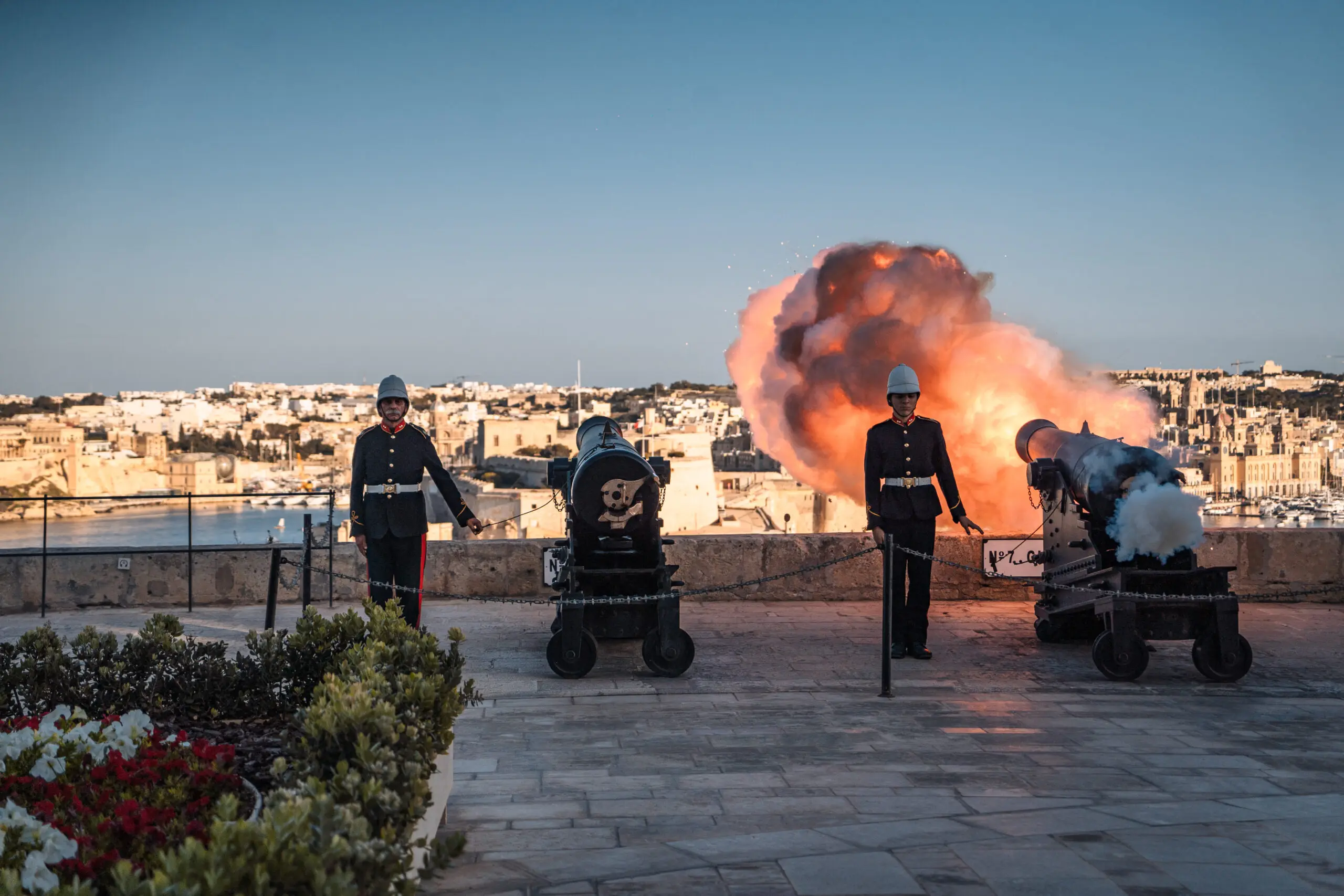 The gun salute at the Saluting Battery: gunners in uniform beside the cannons as one fires over the Grand Harbour, Birgu across the water