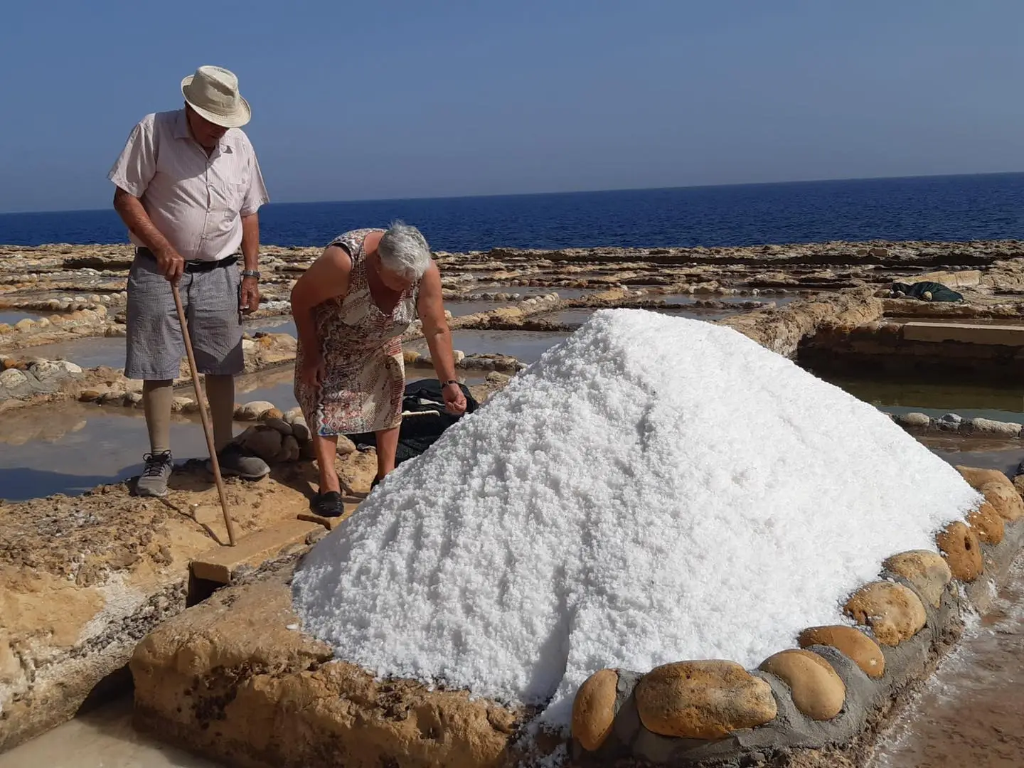 Two salt farmers beside a heap of freshly harvested salt, the pans and the sea behind them