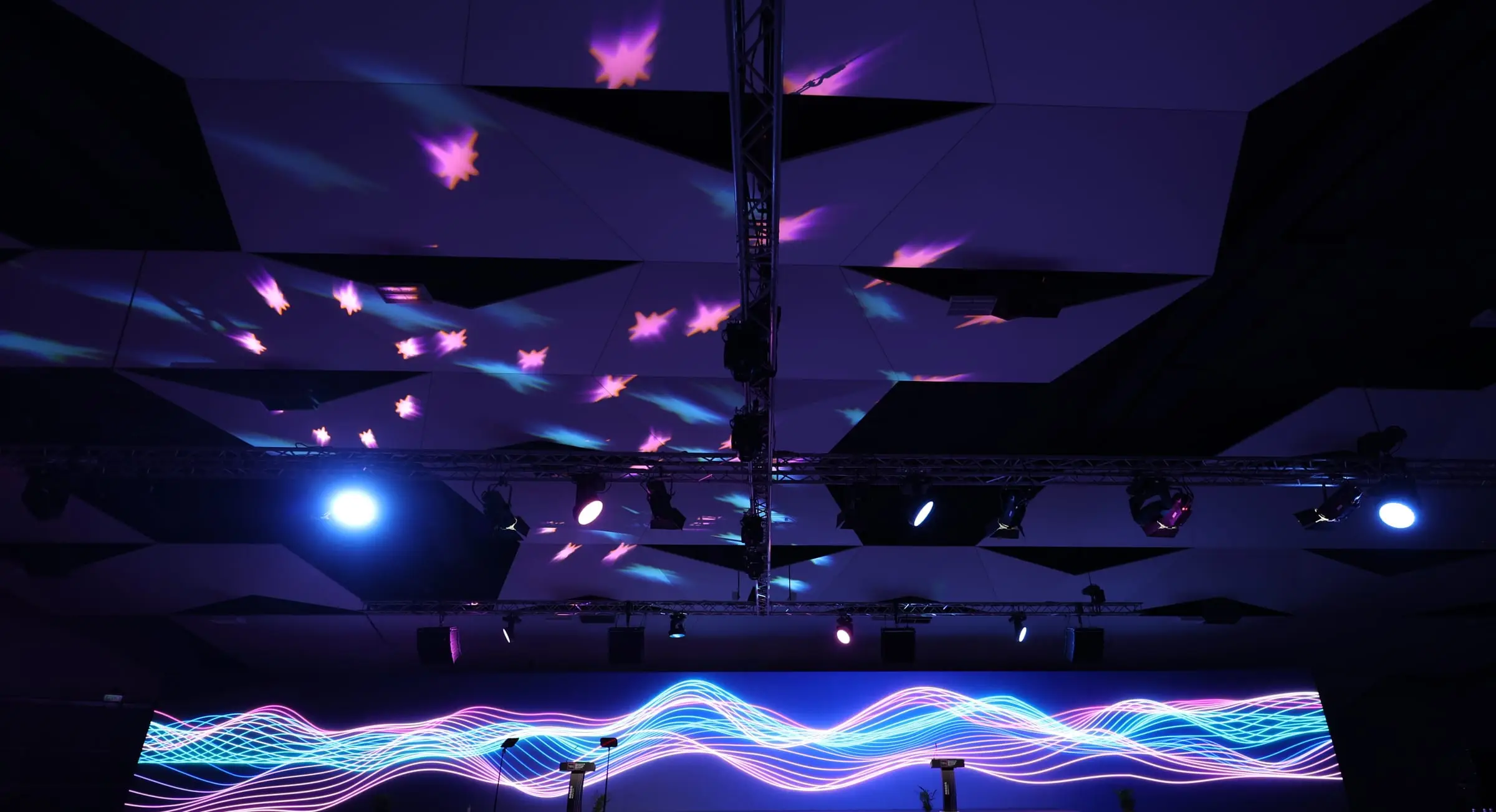 A darkened arena with star-shaped lights projected across its angled ceiling and a long LED wall showing waves