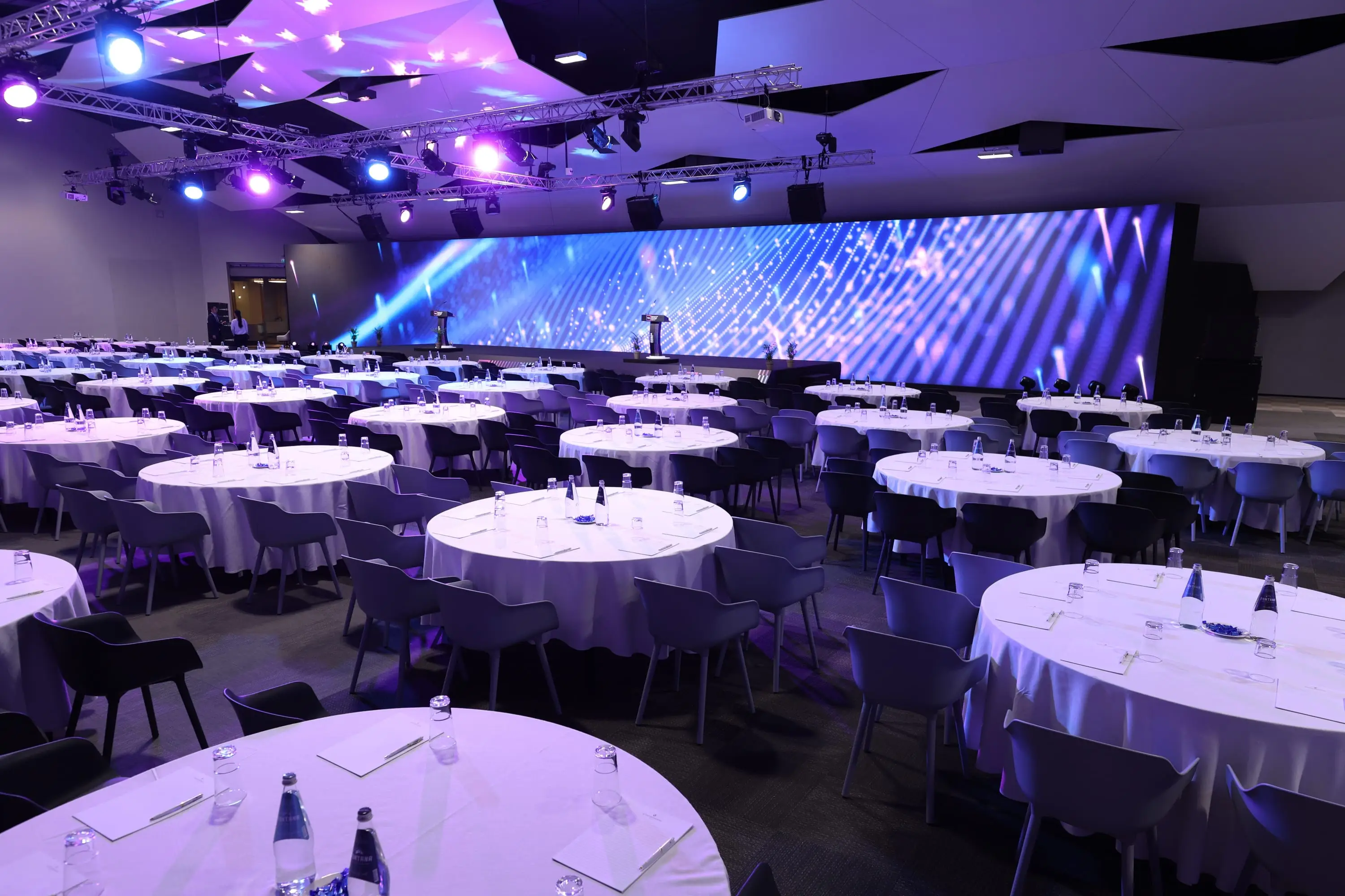 The arena set in rounds: white tablecloths and dark chairs beneath the lighting rig, a full-width LED wall behind the stage