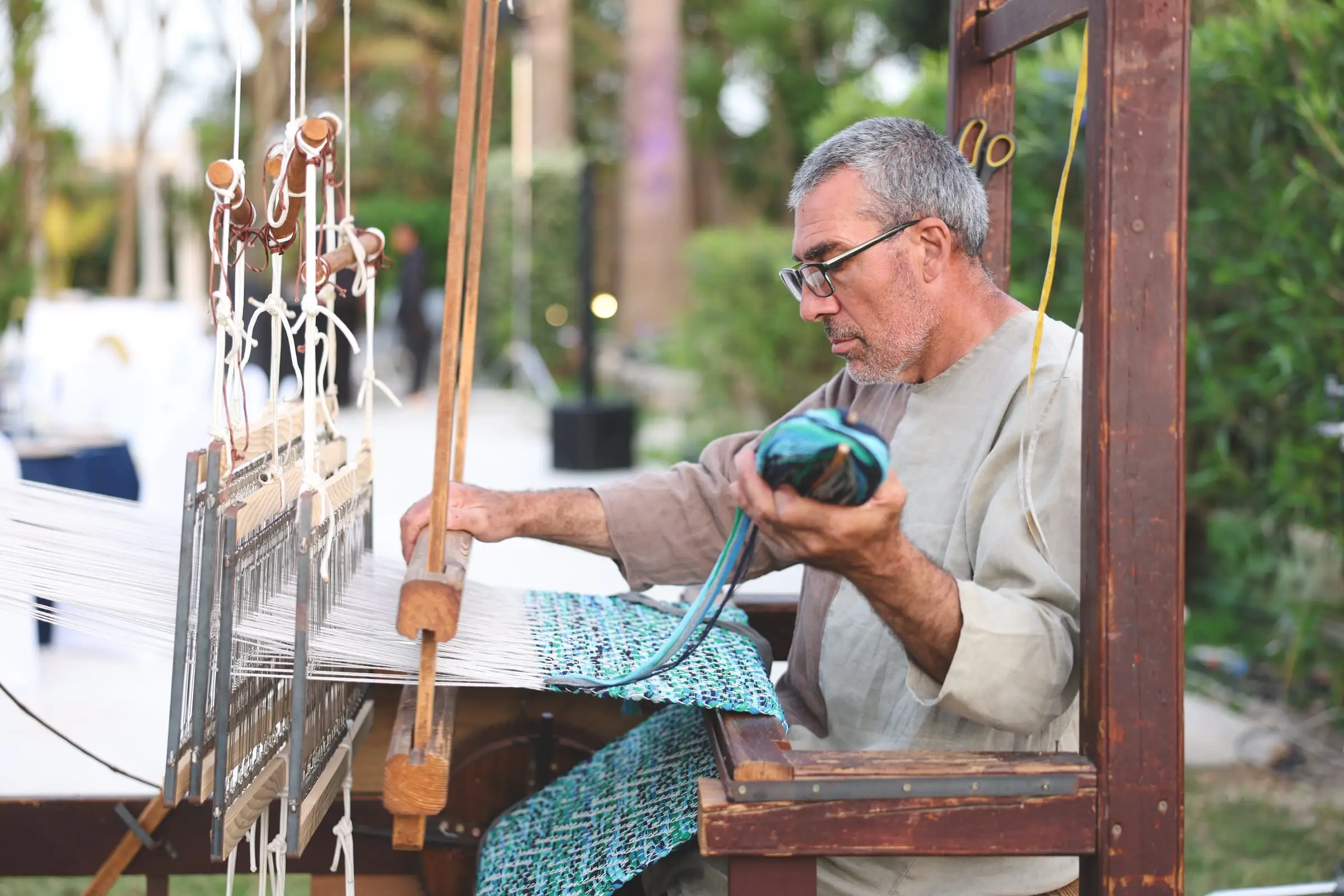 A weaver throwing the shuttle on a wooden hand loom, a blue and green cloth growing on the warp