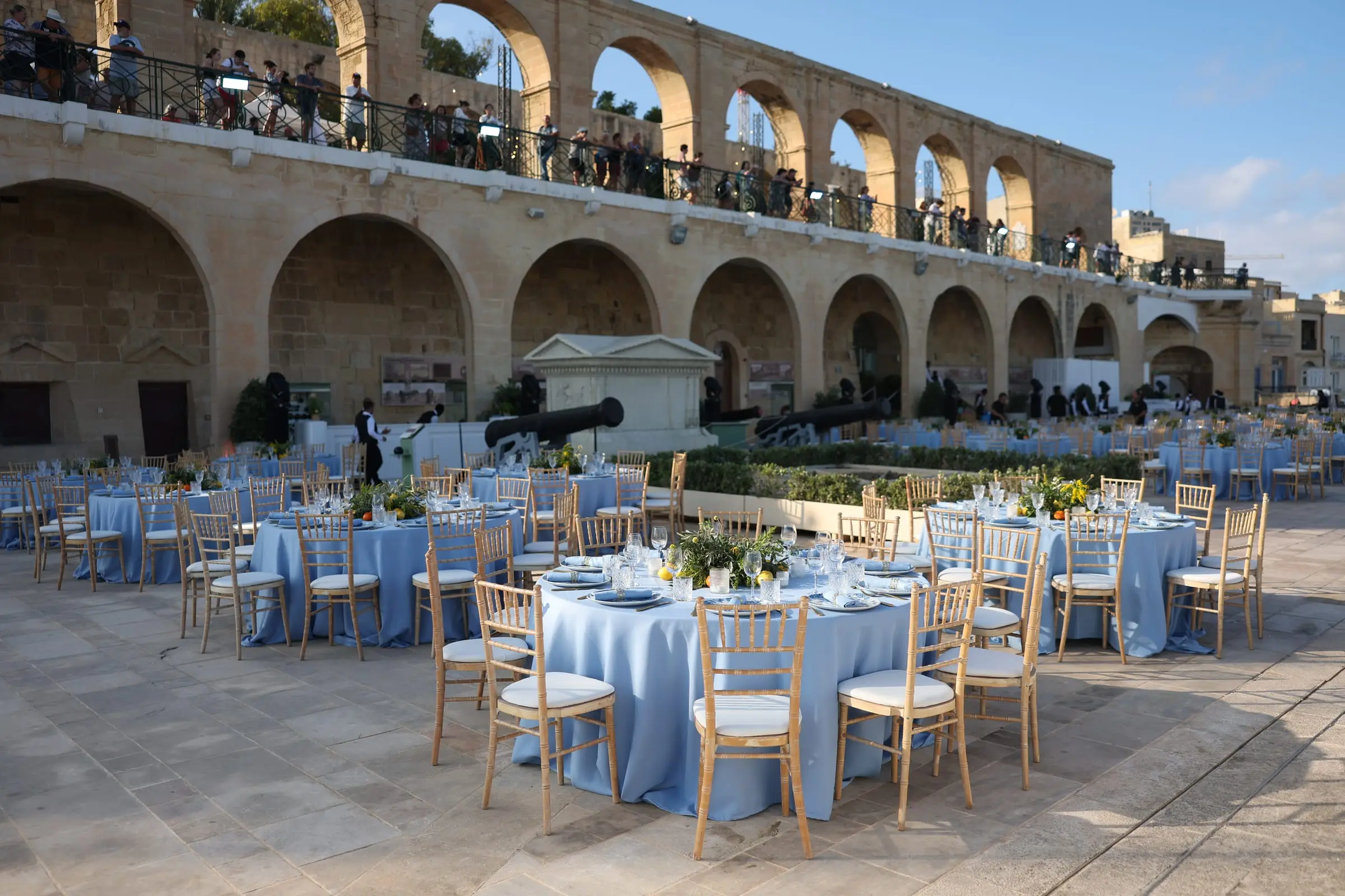 Round tables in pale blue linen set beneath the arches of the Upper Barrakka Gardens, with the public terrace above