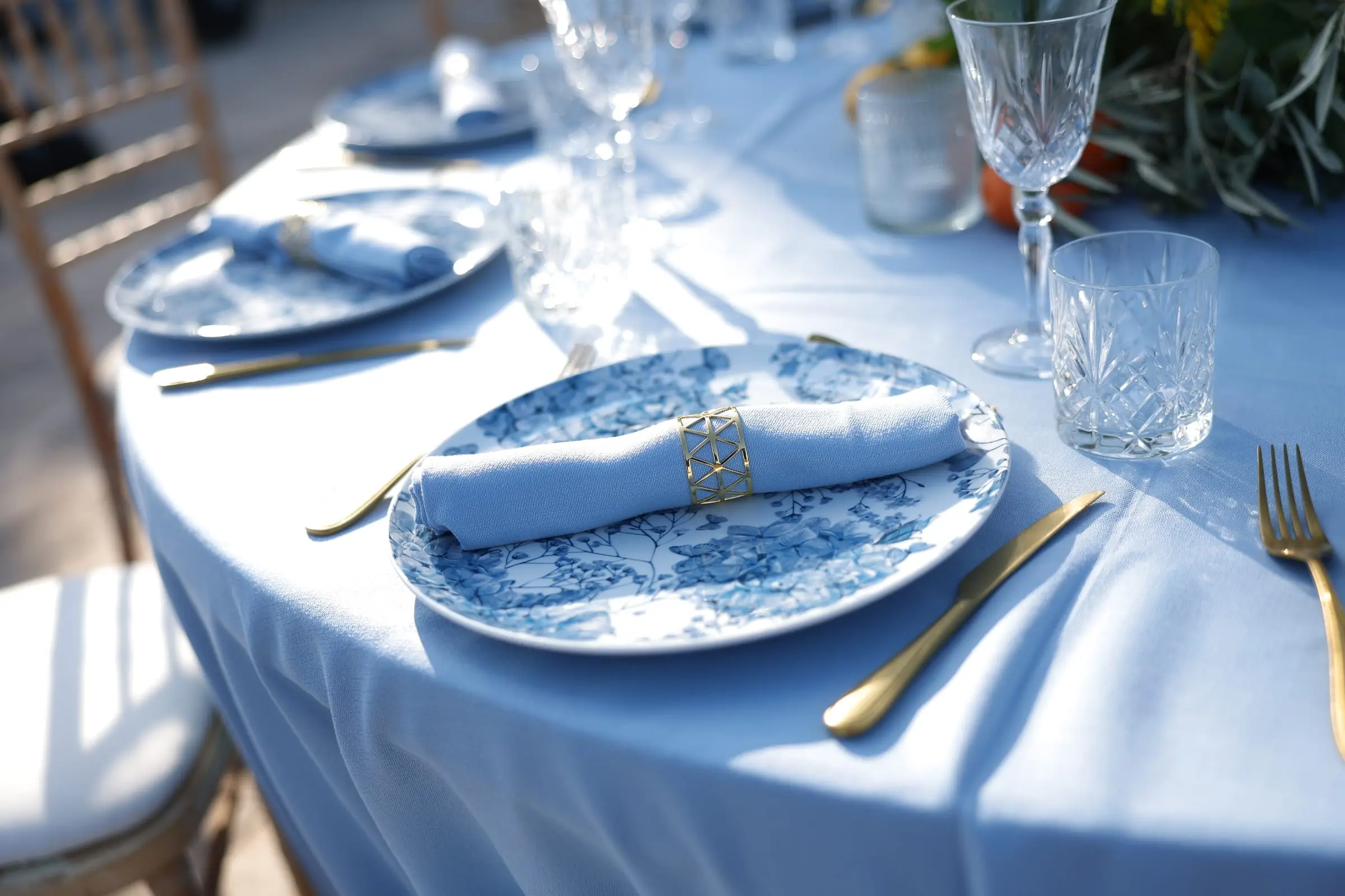 A place setting in pale blue: a floral-patterned plate, a gold napkin ring and gold cutlery beside cut glassware