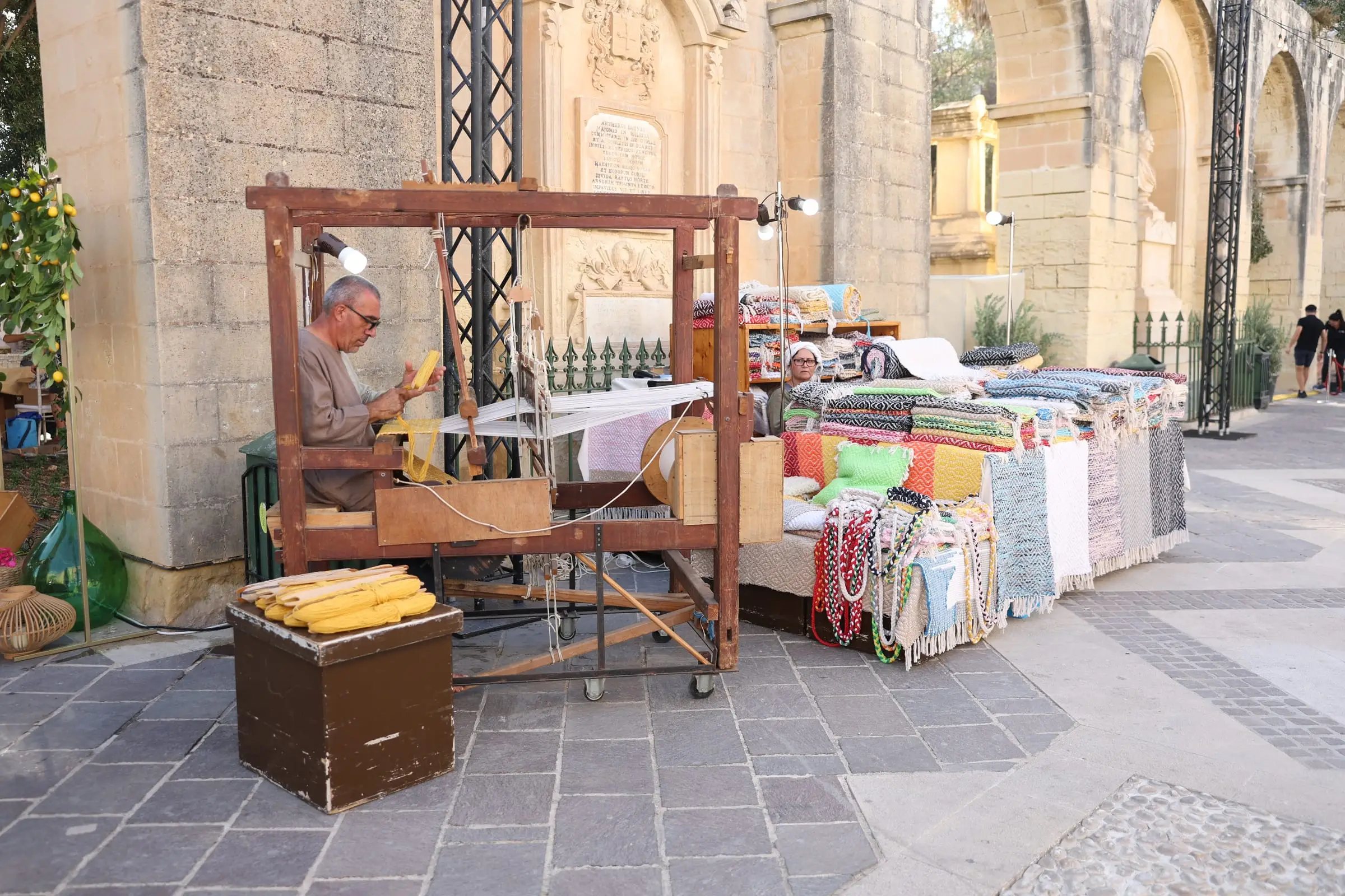 A Maltese artisan weaving at a traditional wooden loom beneath the arches of the Upper Barrakka Gardens