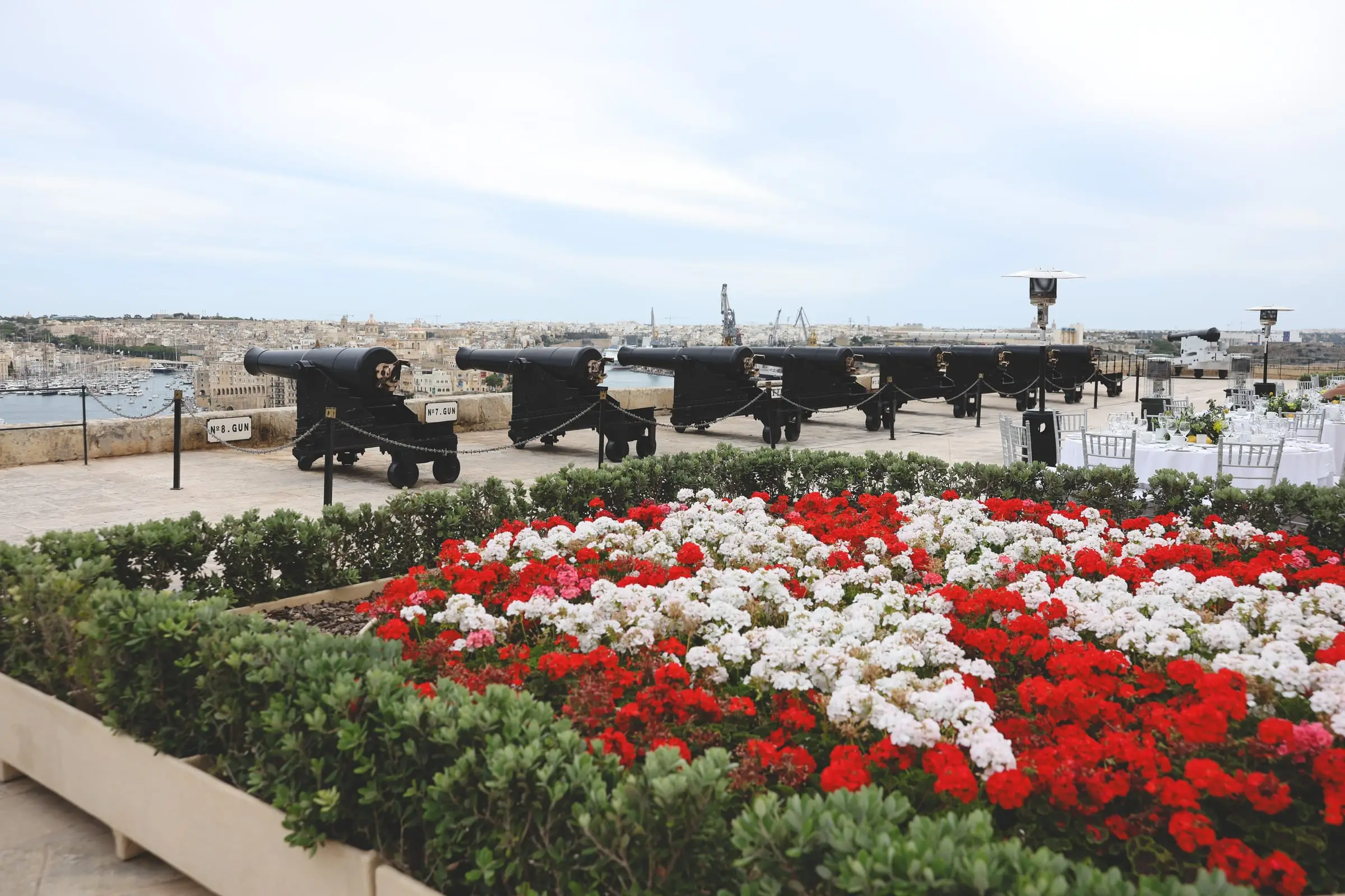 A bed of red and white geraniums planted as a Maltese cross, the line of cannons and the harbour behind