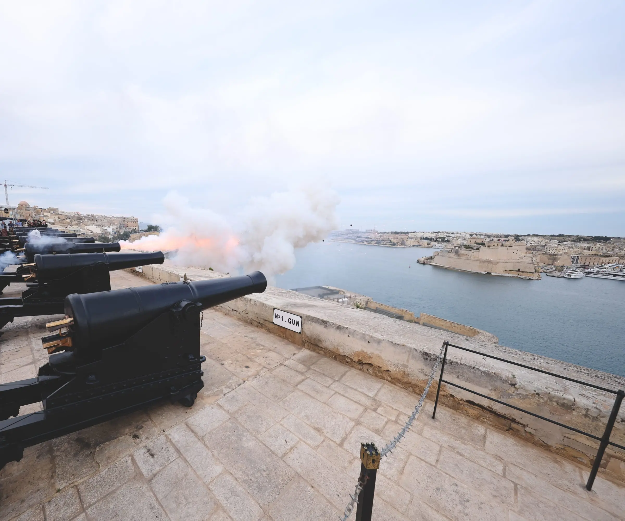 The cannons of the Saluting Battery fire in line along the parapet, smoke rolling out over the Grand Harbour