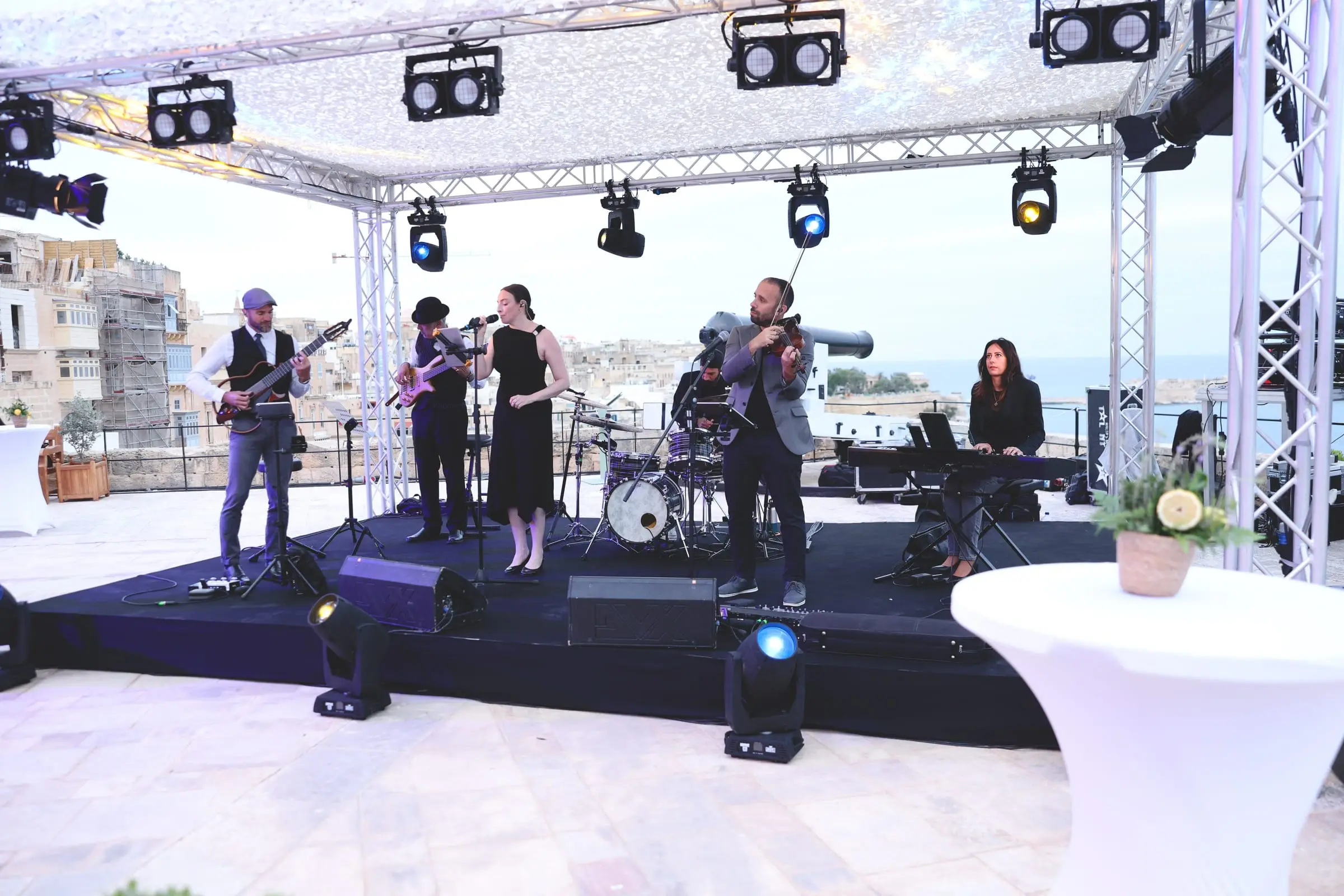 A six-piece band with guitars, a singer, violin, drums and keys plays under a lit truss stage on the Saluting Battery, with Valletta's limestone houses behind