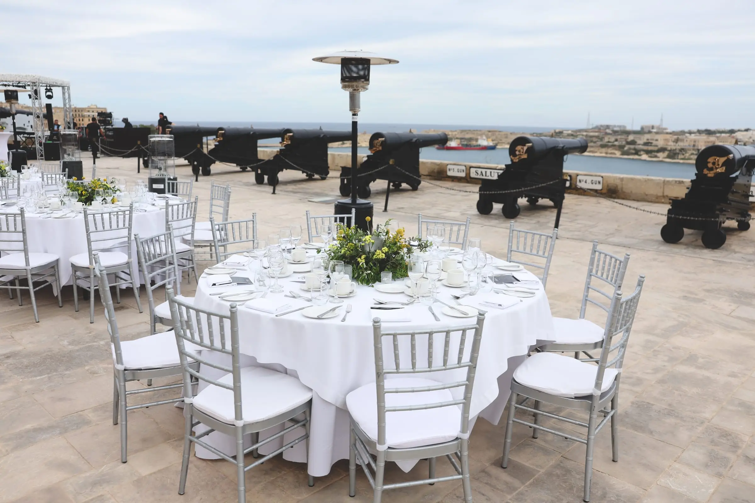 A round table laid in white with silver chairs and a green and yellow centrepiece, the black cannons of the Saluting Battery behind it and the harbour beyond