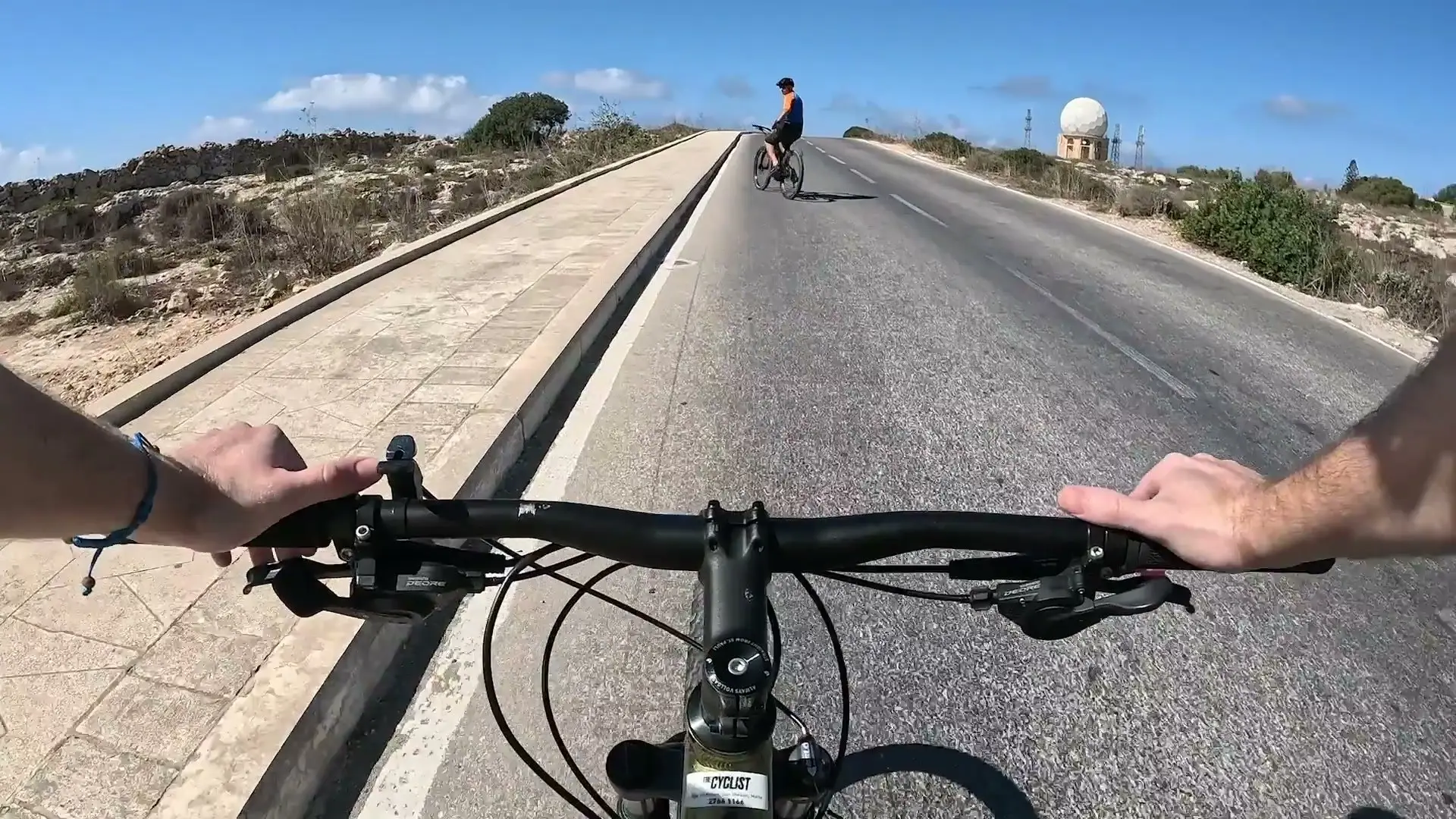 The view over an e-bike's handlebars on a coast road, a cyclist ahead and a white radar dome on the ridge