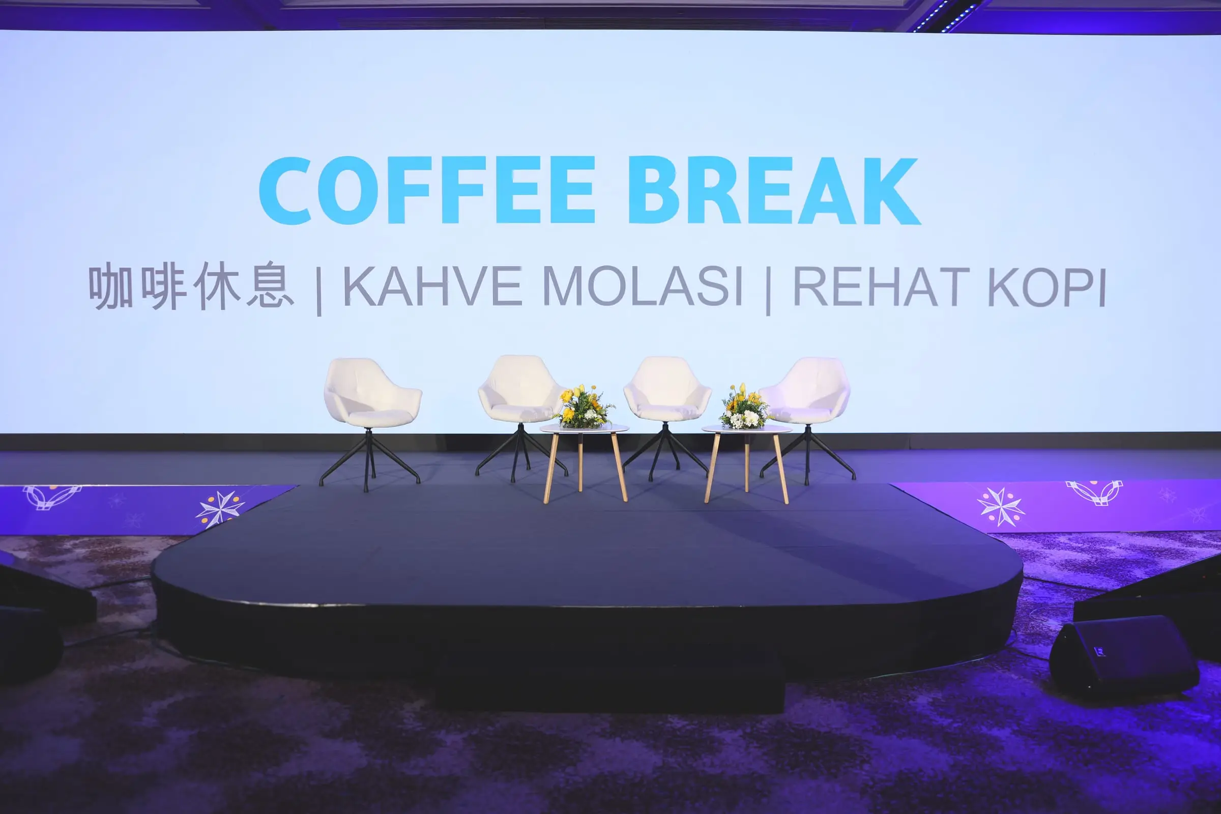The plenary stage between sessions: four white armchairs and two side tables with yellow flowers, the screen behind reading Coffee Break in English, Chinese, Turkish and Malay