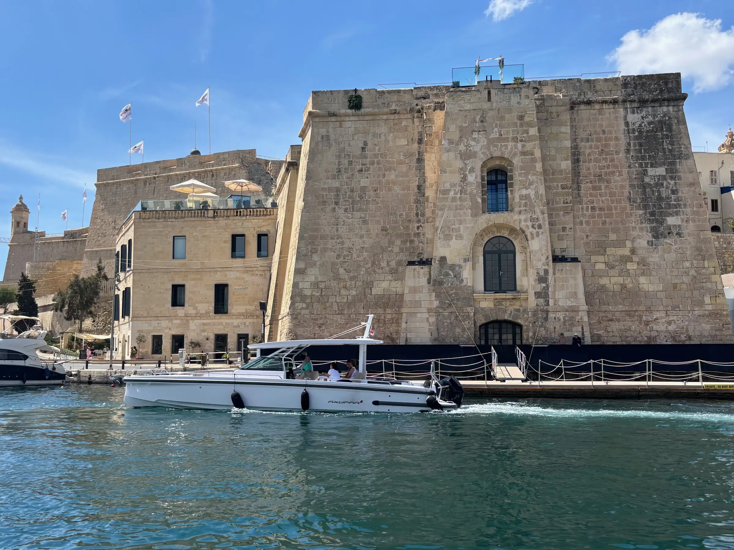 A white day boat with guests aboard crossing the Grand Harbour below a limestone bastion