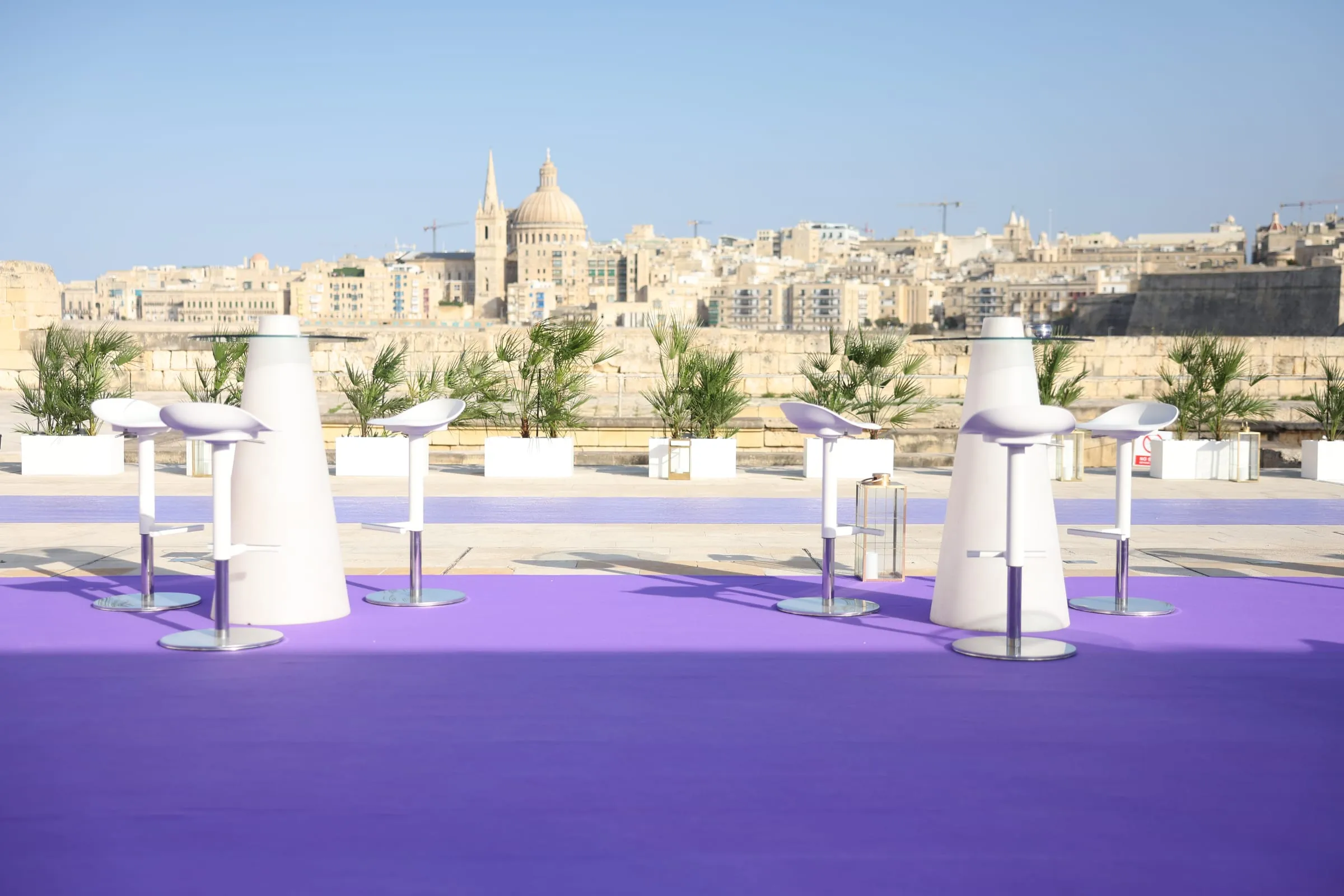 White bar tables on a purple carpet along Fort Manoel's bastion, Valletta's skyline across the harbour