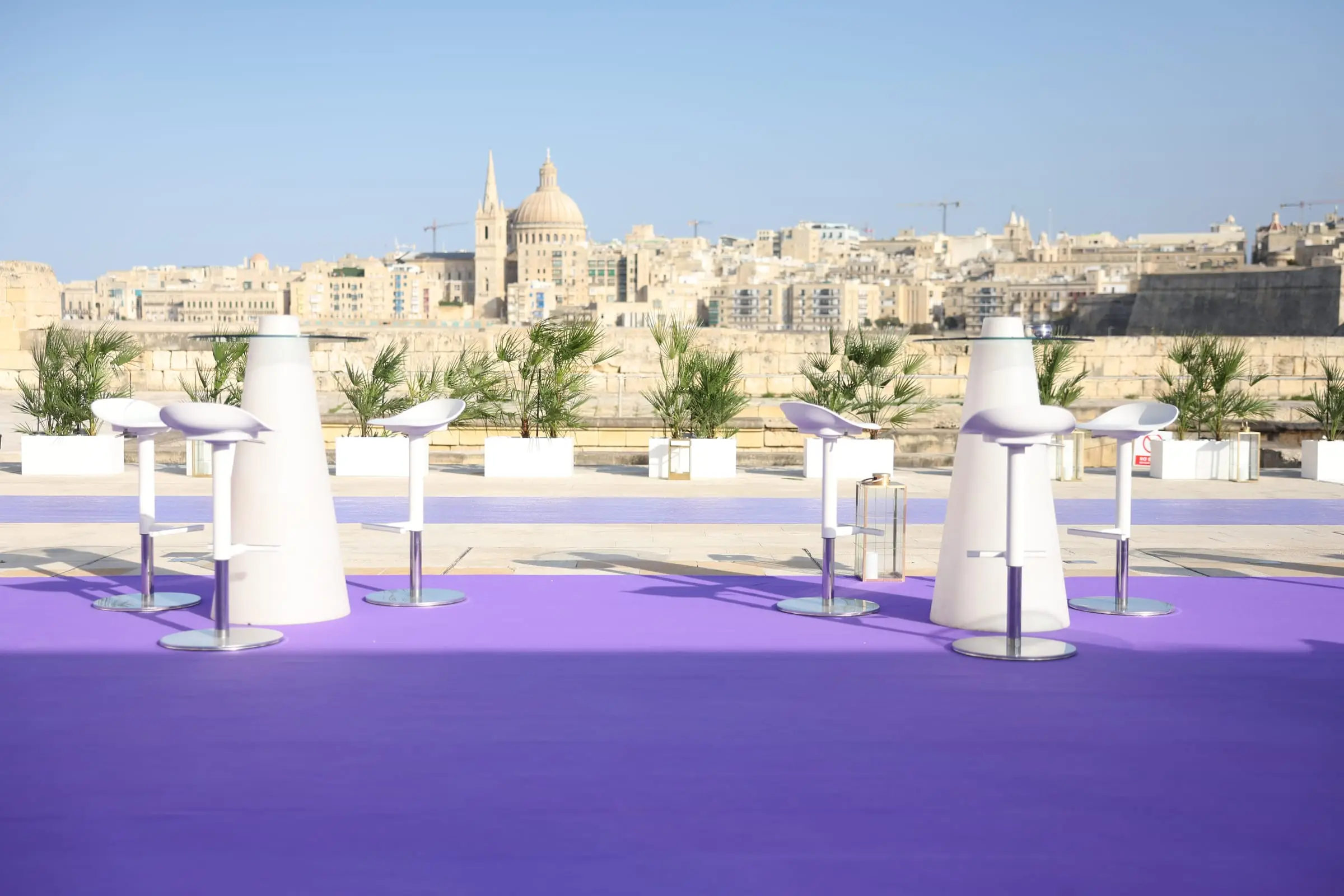 White bar tables on a purple carpet along Fort Manoel's bastion, Valletta's skyline across the harbour