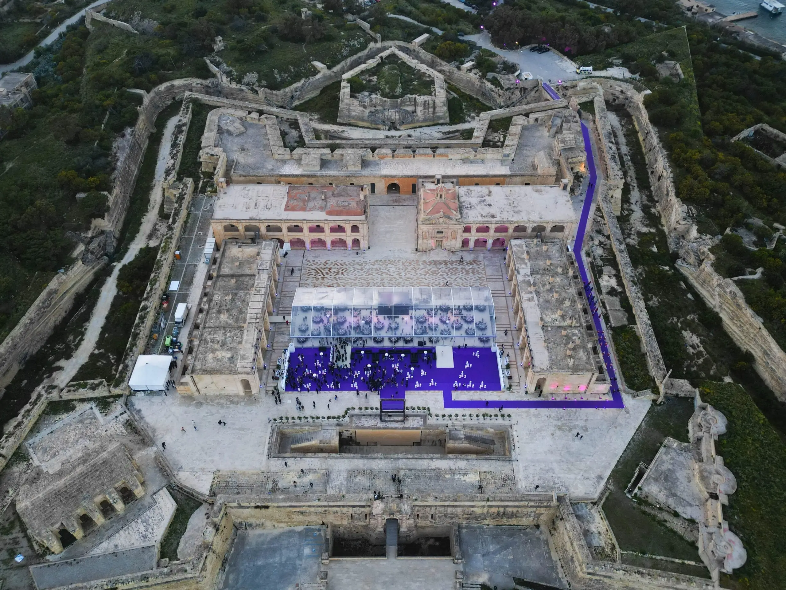 Fort Manoel from the air at dusk: the star fort's walls and parade ground, a clear-roofed marquee laid for dinner, and a purple carpet running from the gate to a reception crowded with guests