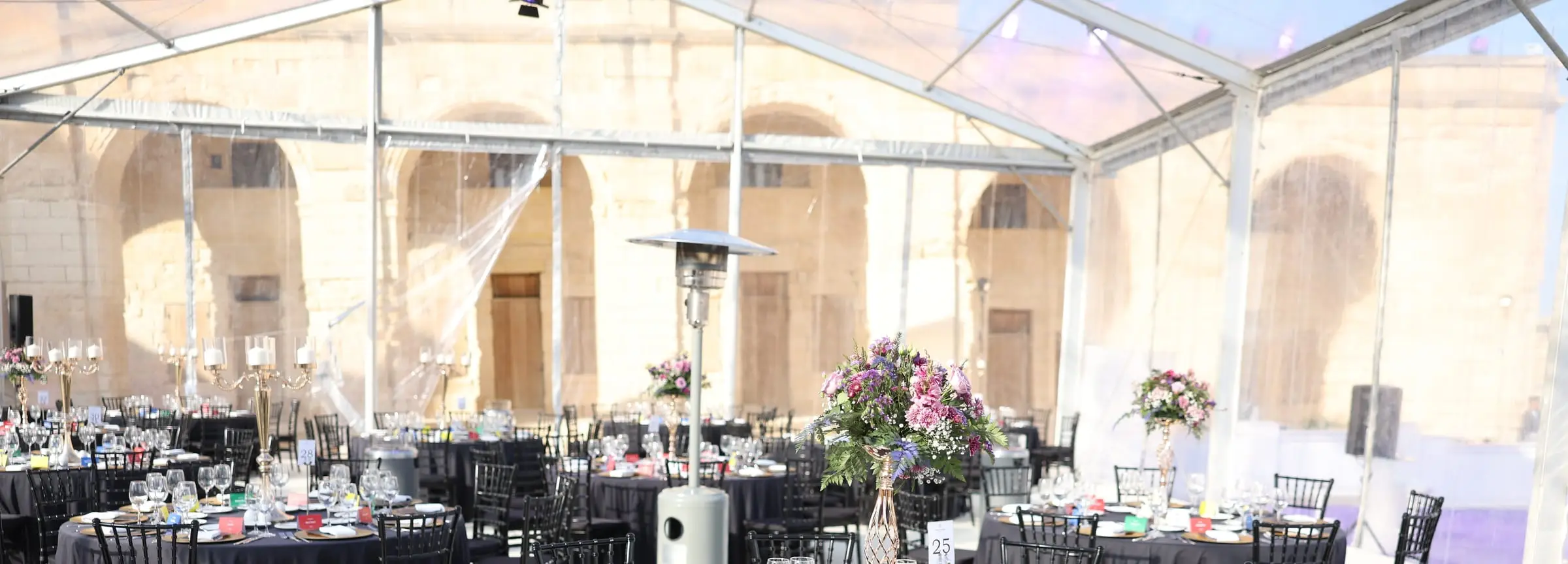A clear-roofed marquee inside Fort Manoel: round tables in black linen with black Chiavari chairs and tall floral centrepieces, the fort's stone arcade behind