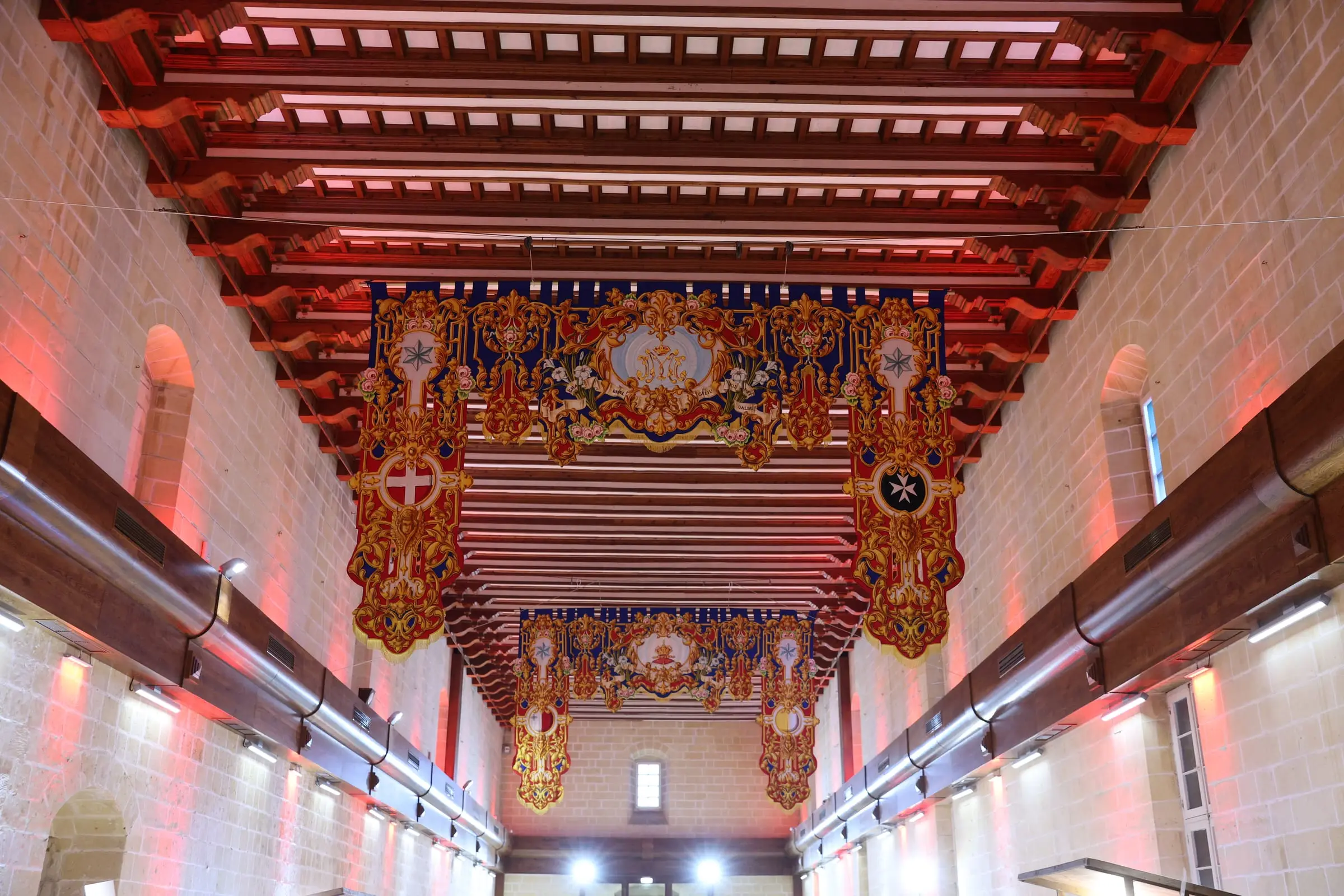 The long hall of the Sacra Infermeria seen from below: a timber-beamed ceiling hung with the Order of St John's red and gold banners, walls washed in red light