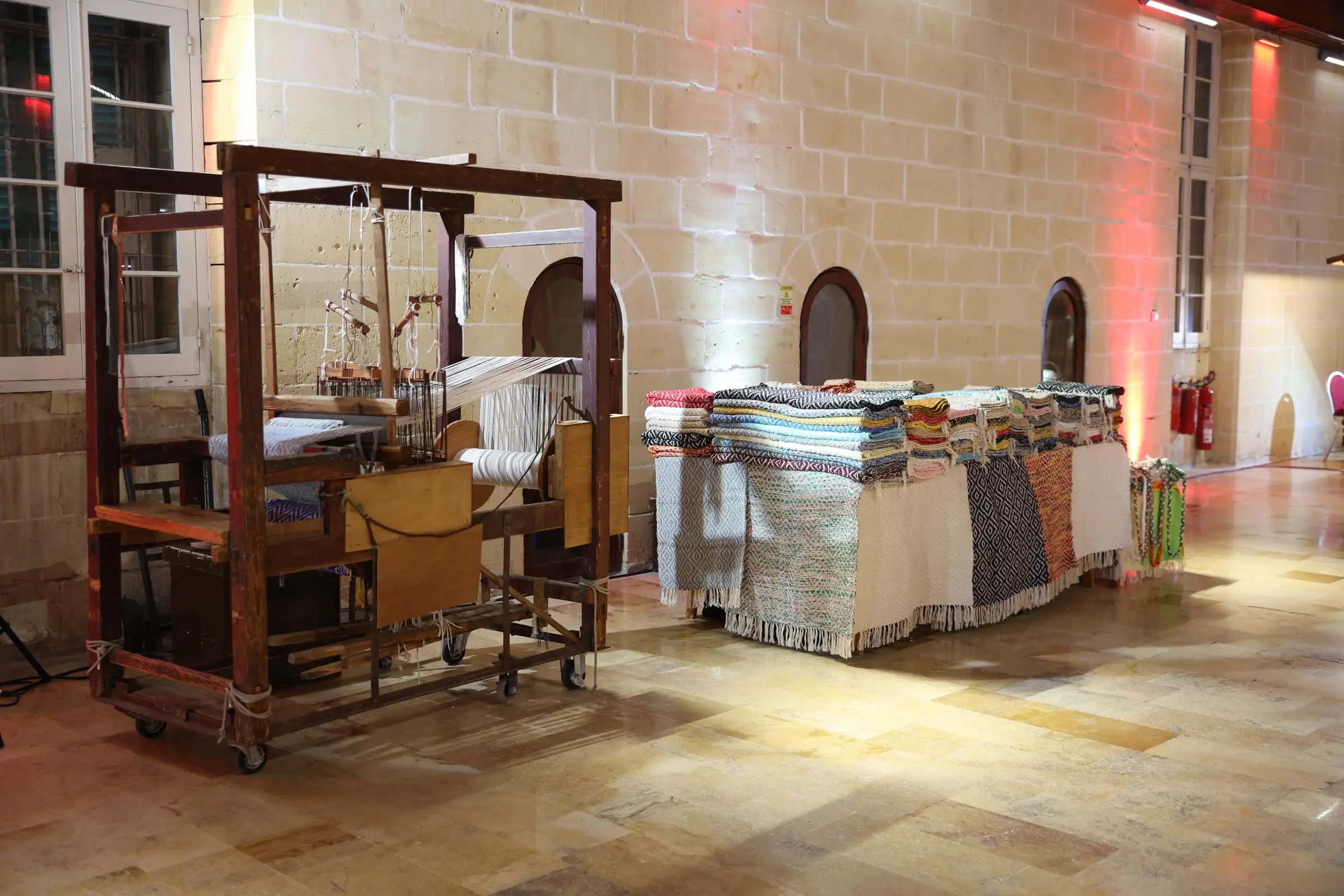 A wooden weaver's loom and stacks of woven textiles set up on the stone floor of the Sacra Infermeria