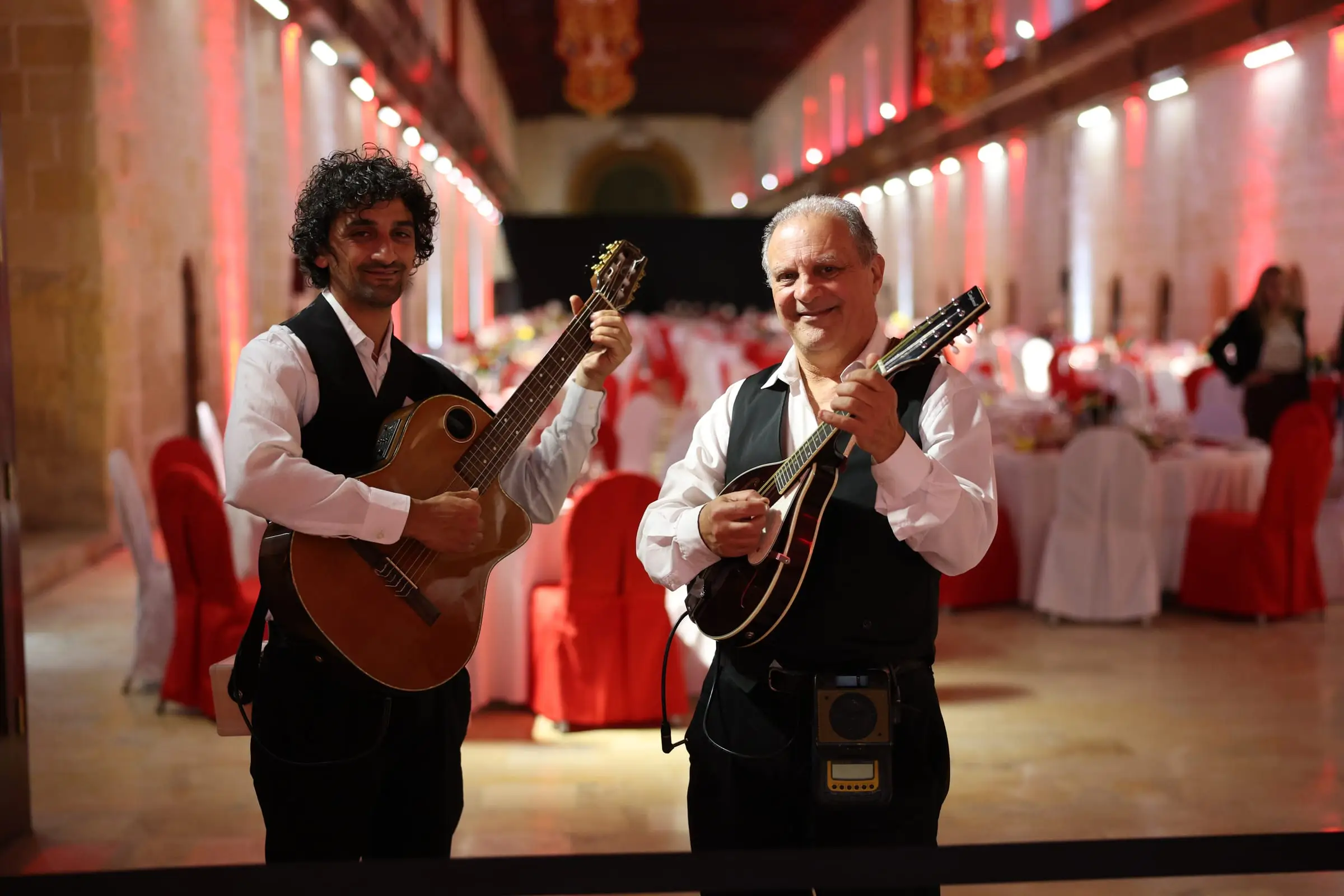 Two musicians in waistcoats, one with a guitar and one with a mandolin, in front of tables dressed in red and white inside the Sacra Infermeria