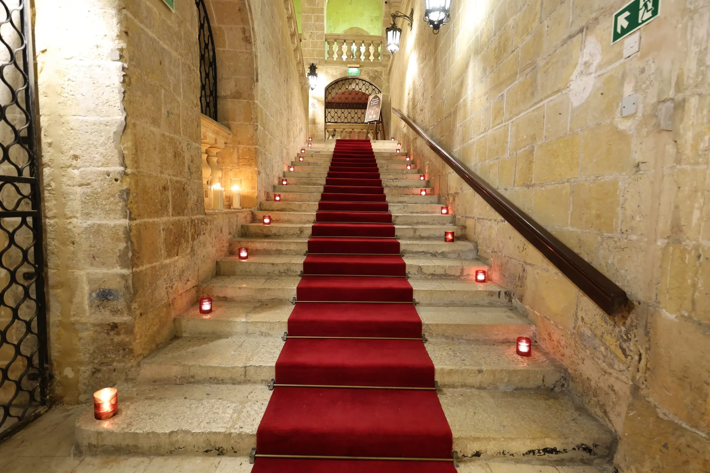 A candle-lined limestone staircase with a red carpet inside the Sacra Infermeria