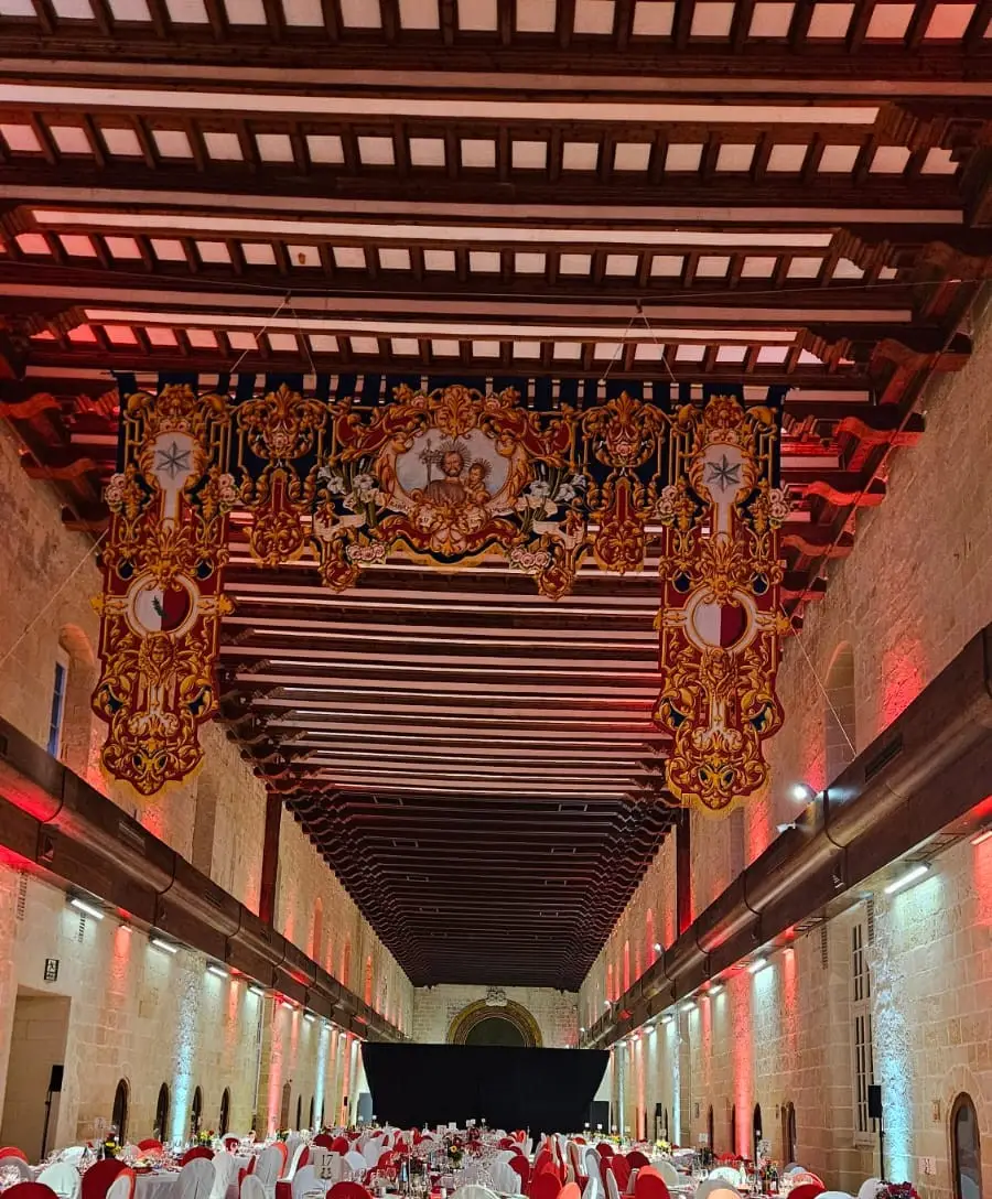 The long ward of the Sacra Infermeria set with round tables in red and white beneath its timber ceiling