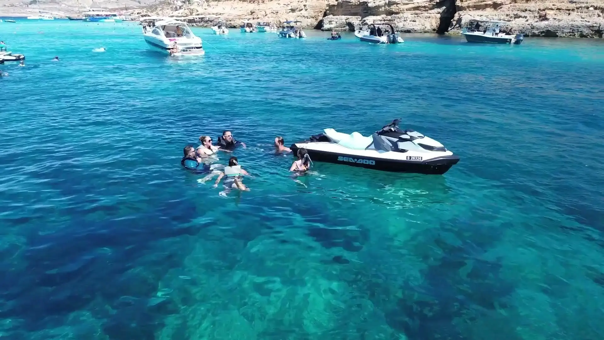 A jet ski on turquoise water seen from above, guests swimming beside it and boats at anchor by the rocks