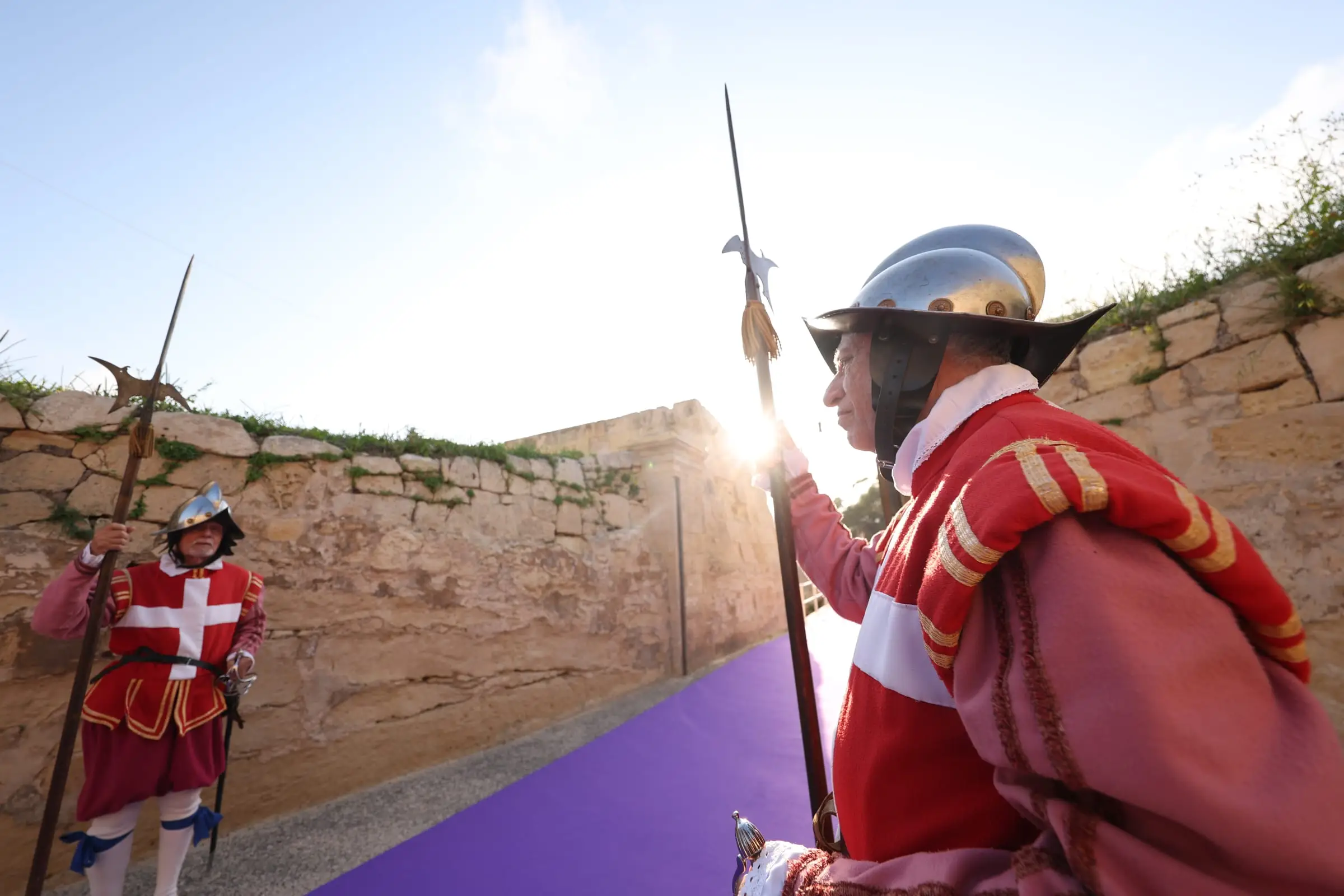 A halberdier of the guard in a red tunic with a white cross and a steel helmet, the low sun behind him on the bastion