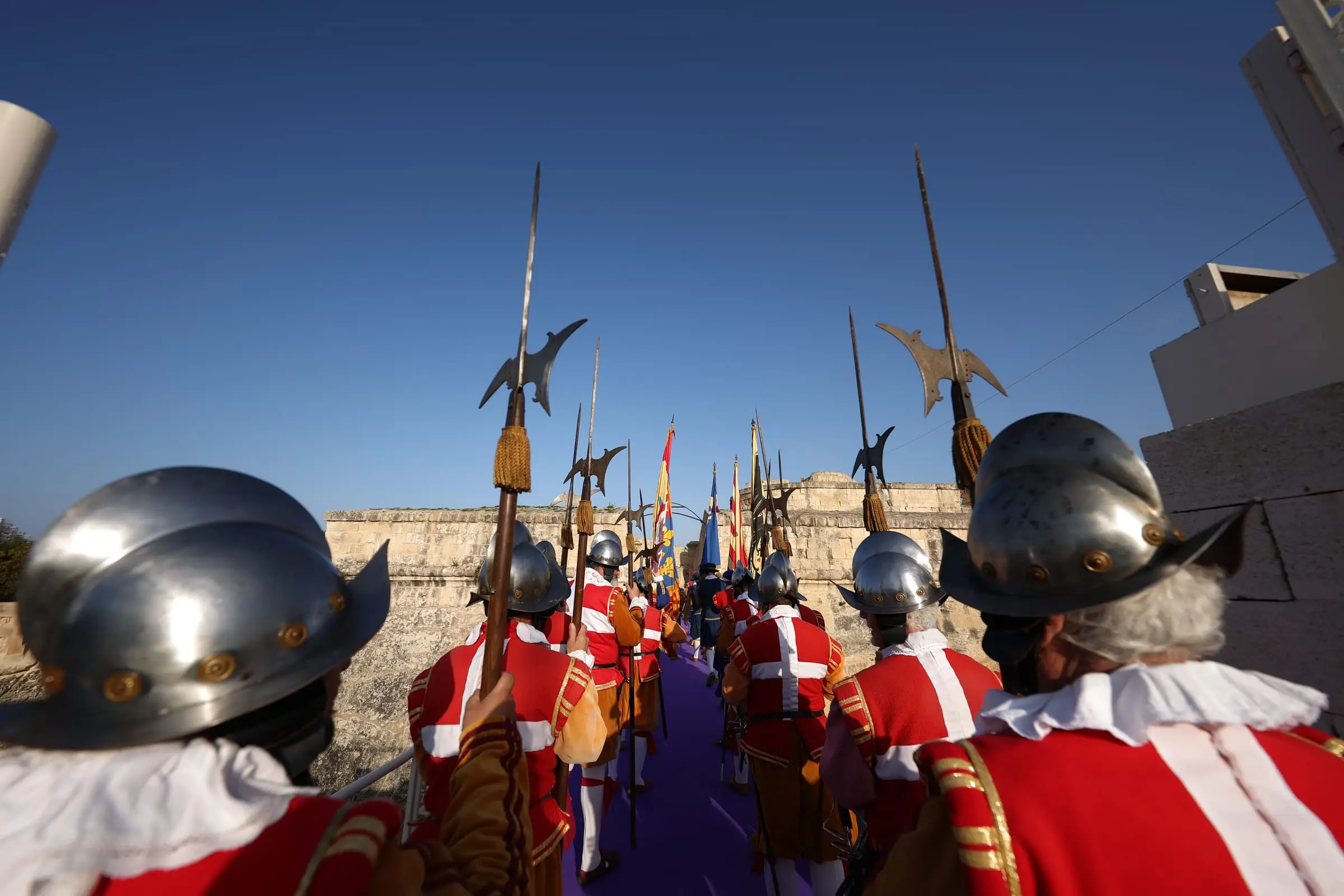 Men of the Knights' guard in red and white tunics and steel helmets, halberds raised, walking away along the purple carpet
