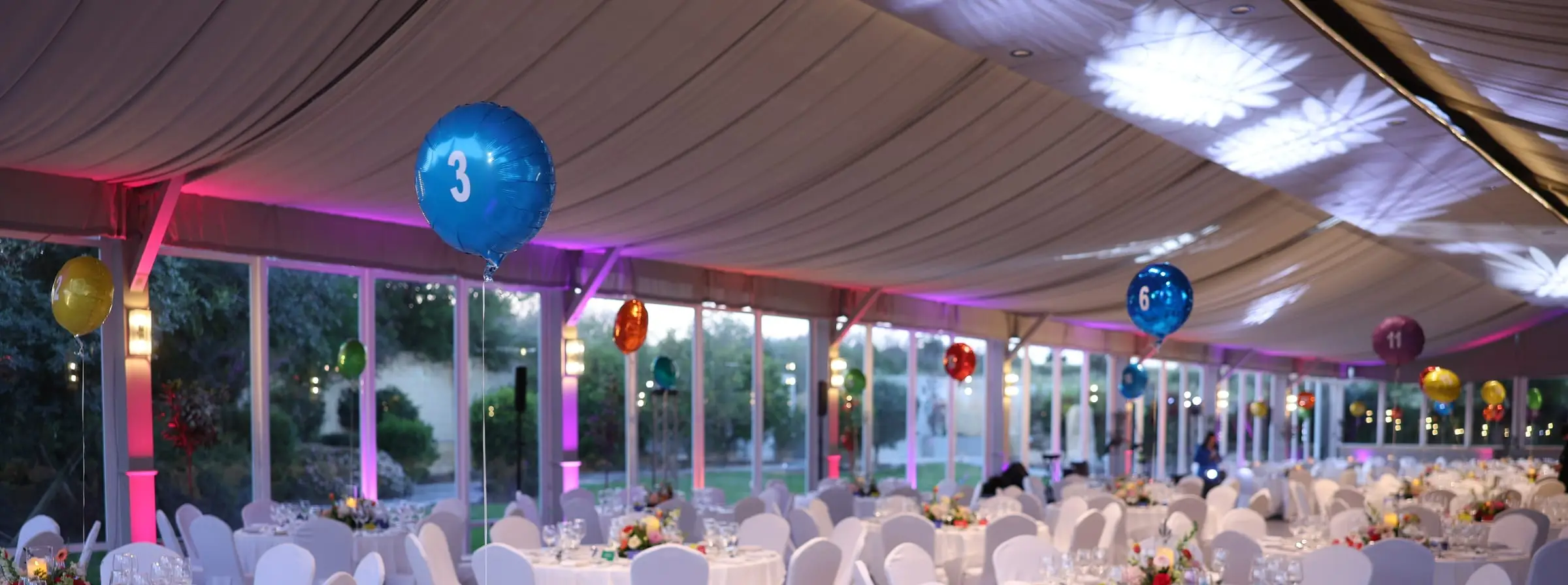 A marquee laid for dinner: white chair covers, floral centrepieces, numbered balloons over the tables, pink uplights and patterns projected on the canopy