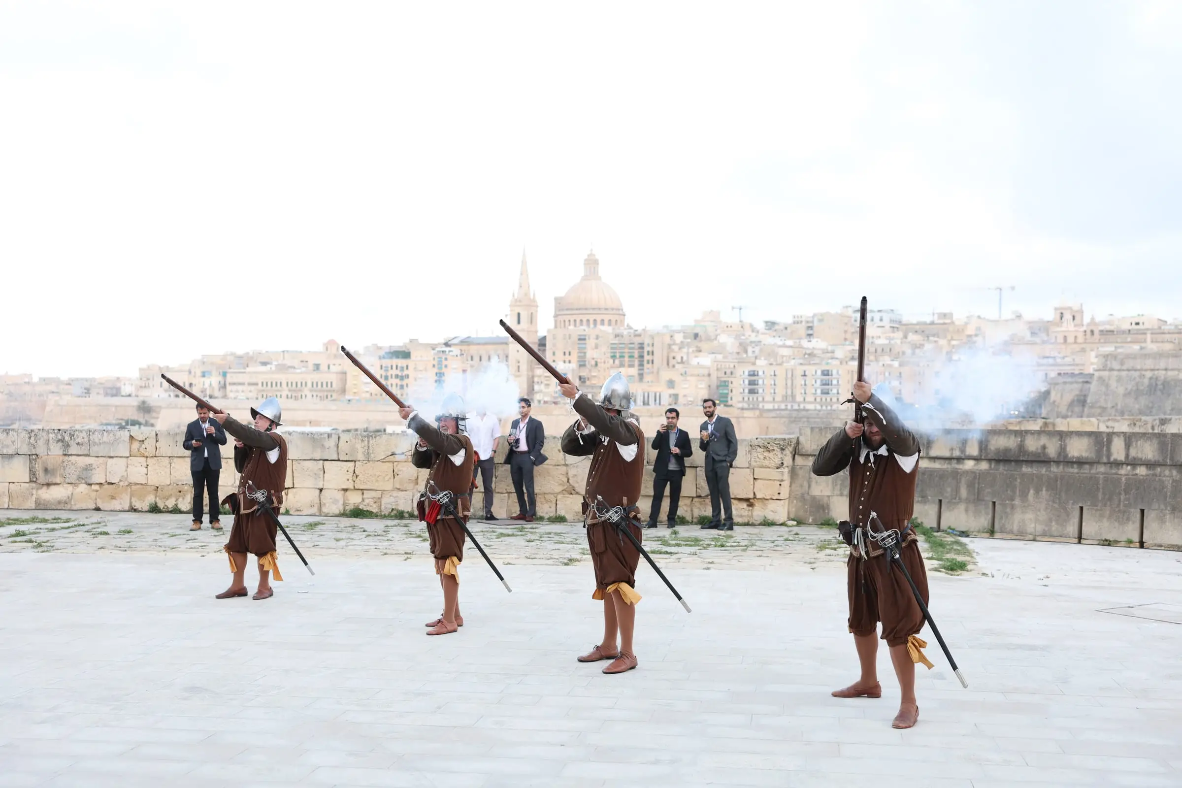 Four musketeers in brown doublets and steel helmets fire a volley on the bastion of Fort Manoel, smoke at the muzzles, the dome and spire of Valletta across the harbour