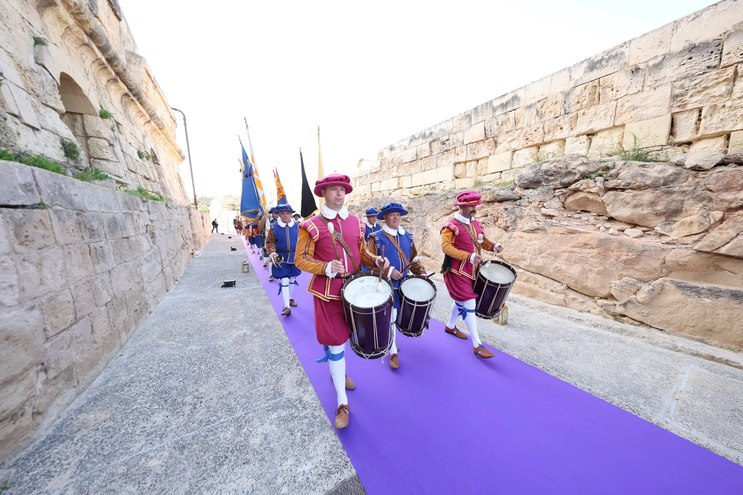 Drummers in magenta and blue Renaissance costume marching along the carpet in the ditch, standard bearers behind