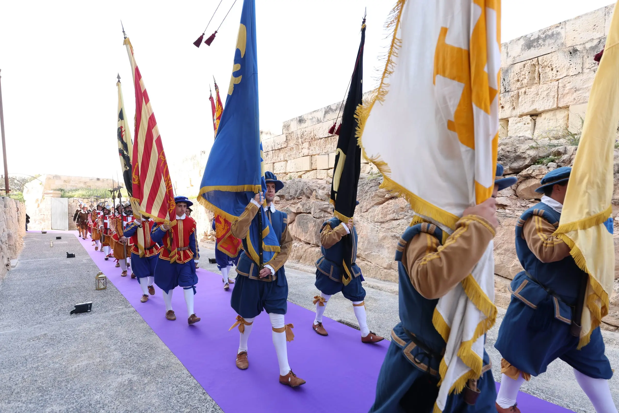 Standard bearers carrying red, gold, blue and black banners along the ditch, the guard following