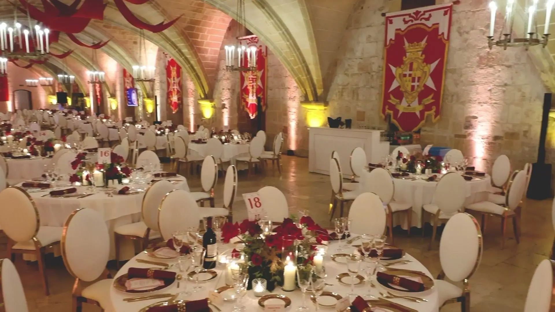 The vaulted hall of the Sacra Infermeria laid with round tables, red roses and candle chandeliers under the Order's banners