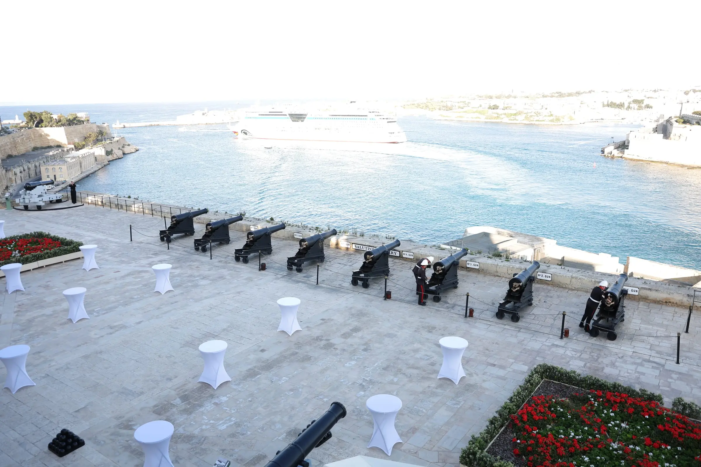 The Saluting Battery's gun platform from above: black cannons along the parapet, white cocktail tables, red geraniums and the Grand Harbour beyond