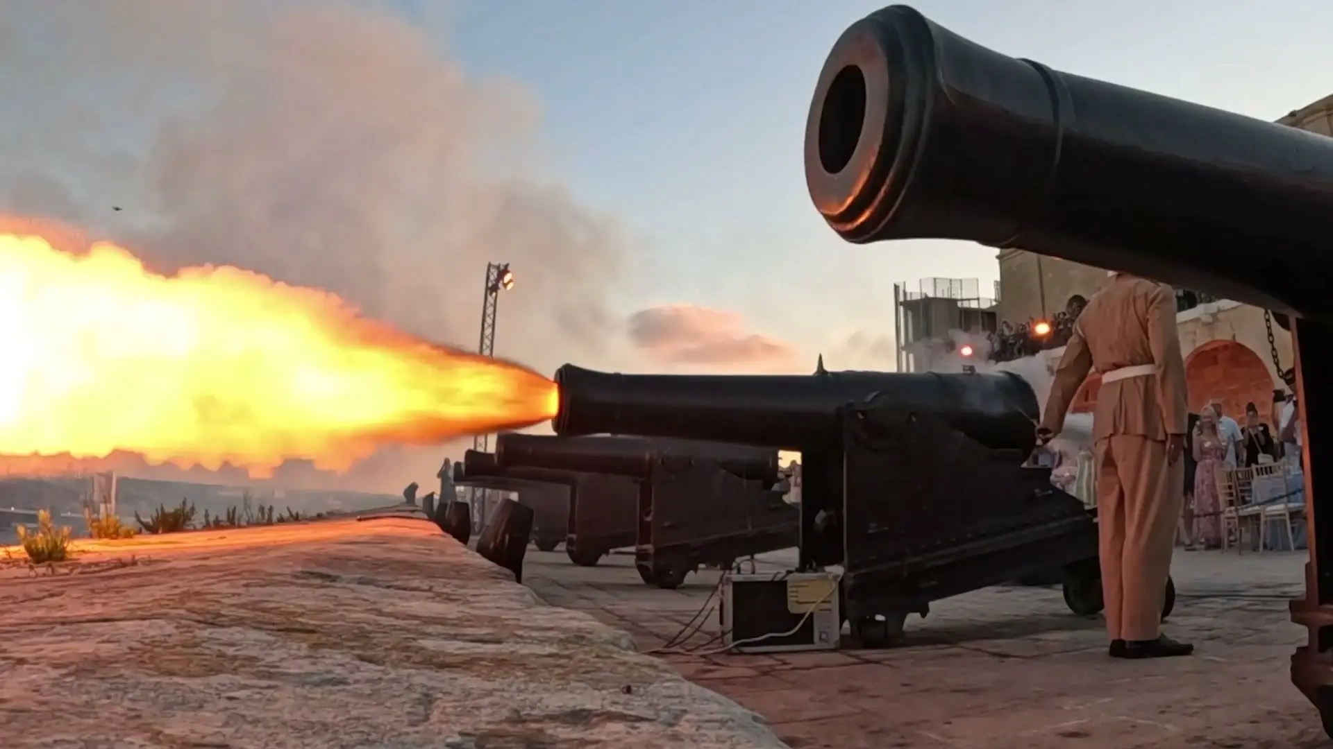 A gunner in period uniform beside a cannon firing over the Grand Harbour at the Saluting Battery