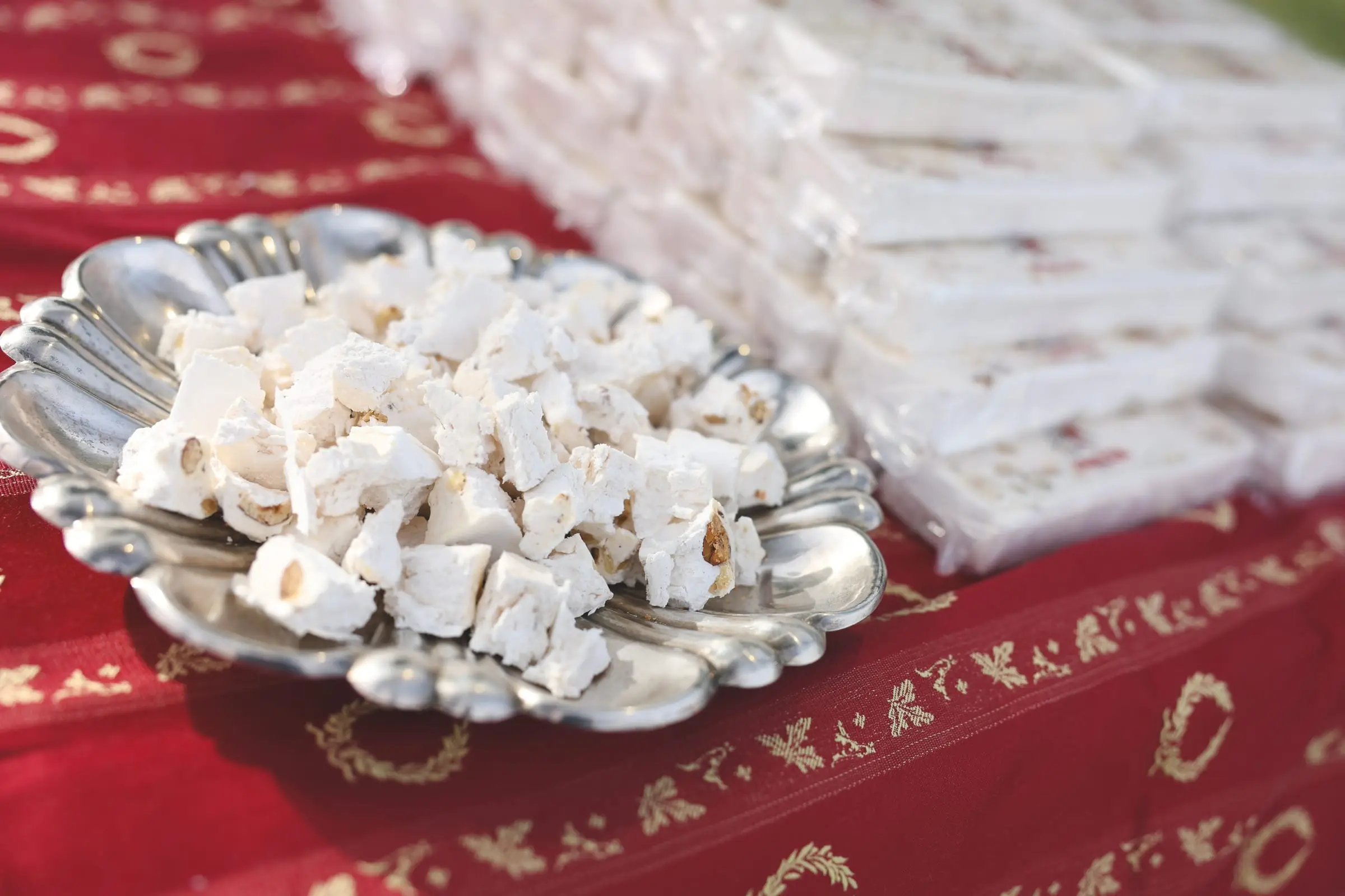 A silver dish of white almond nougat beside wrapped bars on a red and gold cloth