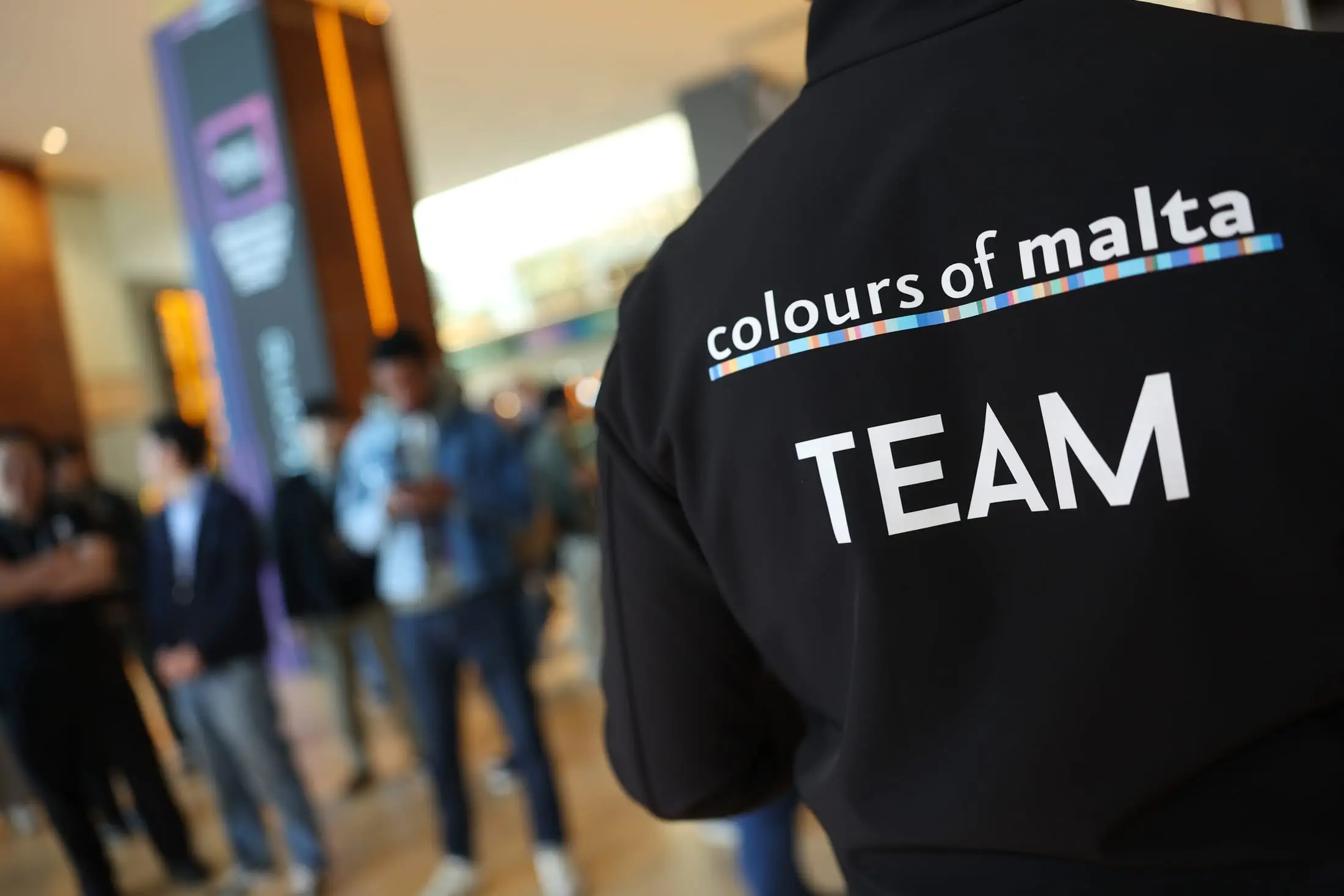The back of a black Colours of Malta team jacket, delegates out of focus in the resort lobby beyond