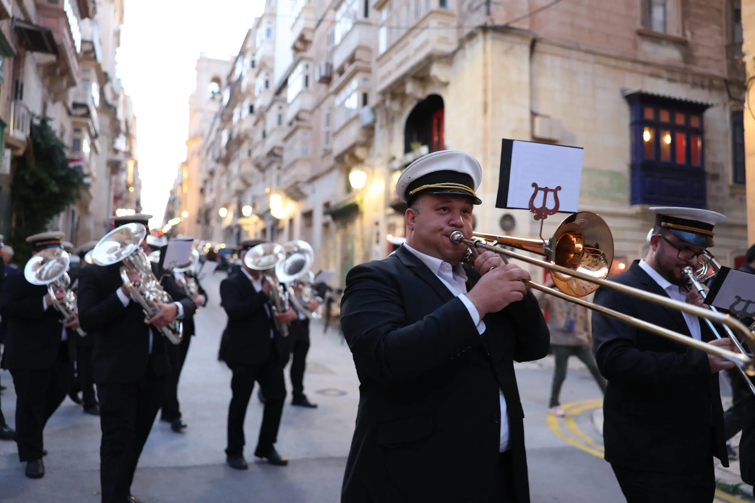 A trombonist in a white-topped cap leads a brass band up a Valletta street of limestone houses and enclosed wooden balconies