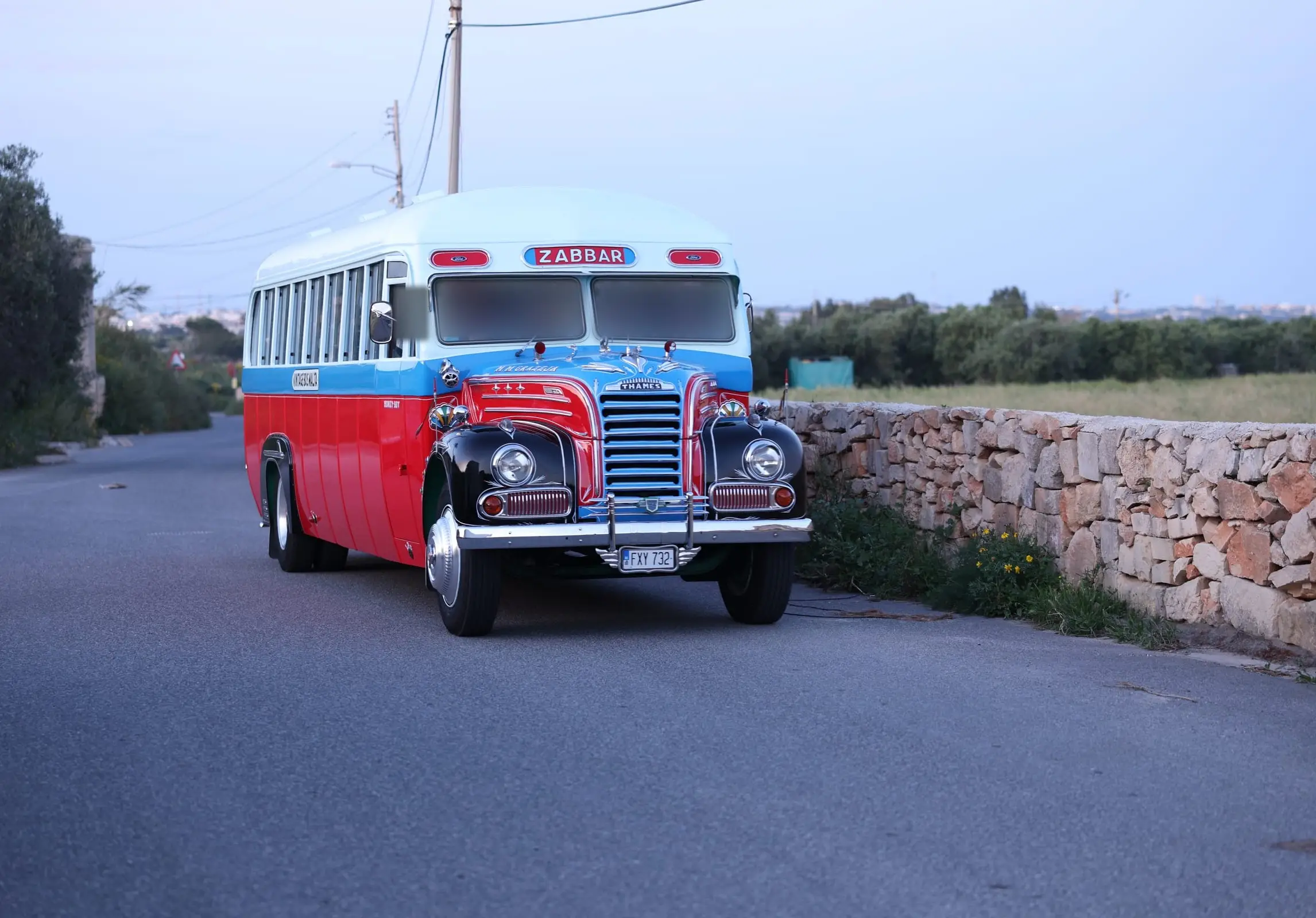 A restored red, cream and blue vintage bus on a country road beside a rubble wall at dusk