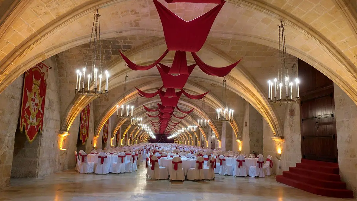 A vaulted hall of the Sacra Infermeria laid with round tables under red drapes and candle chandeliers